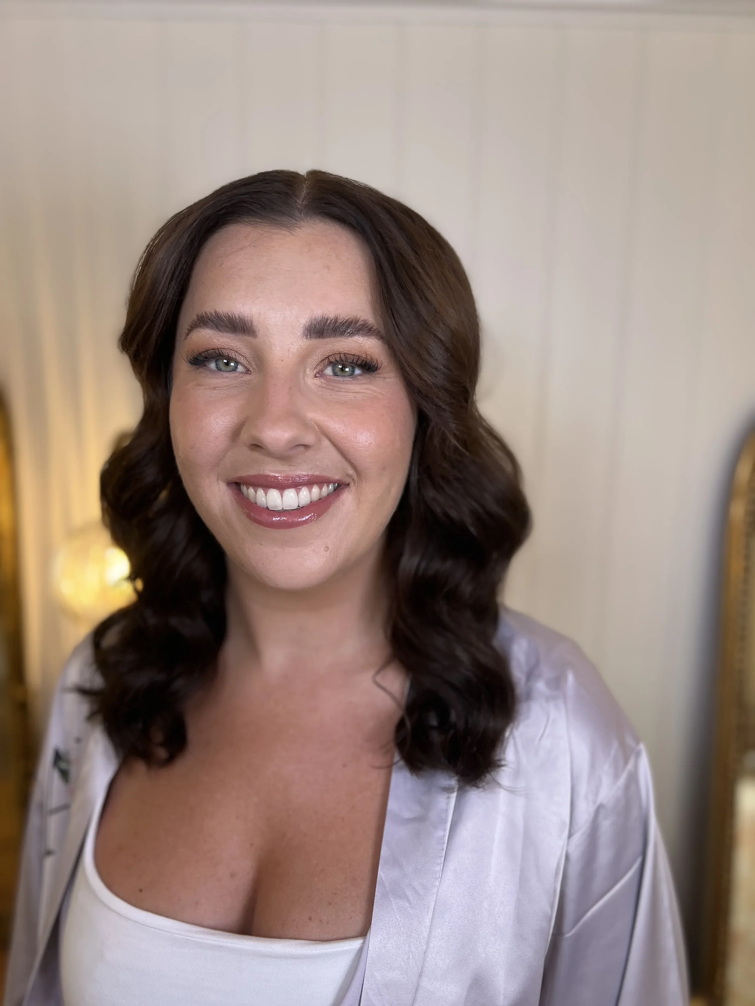 A smiling woman with shoulder-length wavy brown hair, wearing a white top and a silky robe, standing indoors with a neutral background.