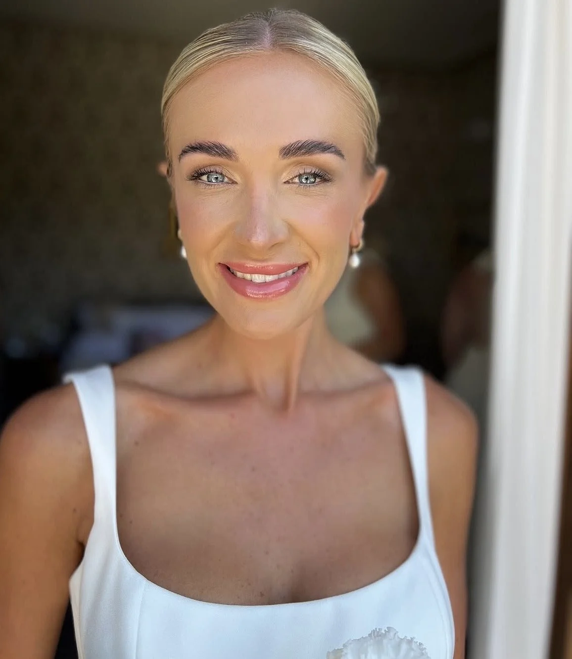 A bride with blonde hair and blue eyes smiling and looking at the camera, wearing a white tank top and pearl earrings, standing near a doorway. She wears natural bridal makeup and a sleek updo.