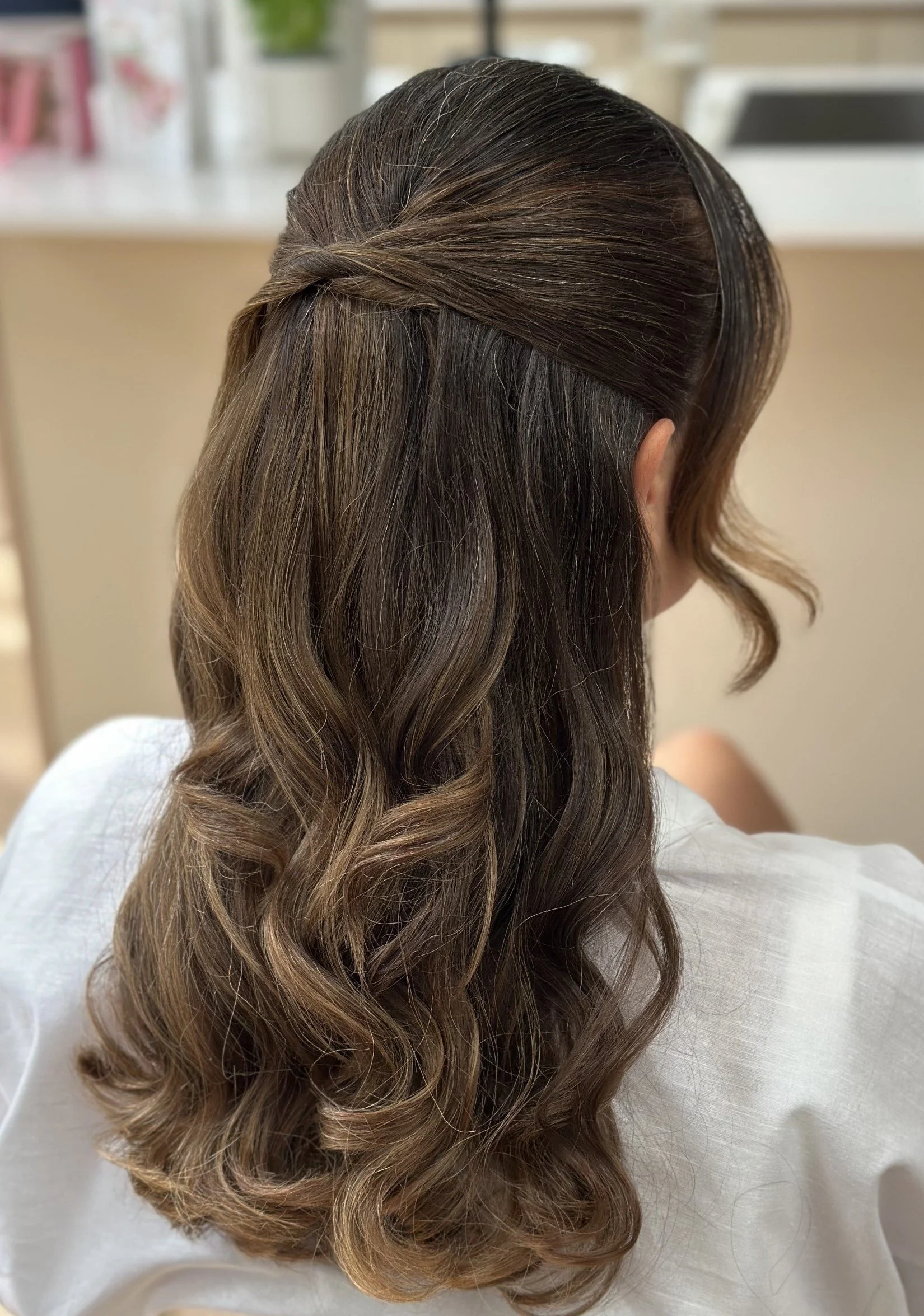 Woman with long, wavy, brown hair styled in loose curls, half-up, wearing a white top.