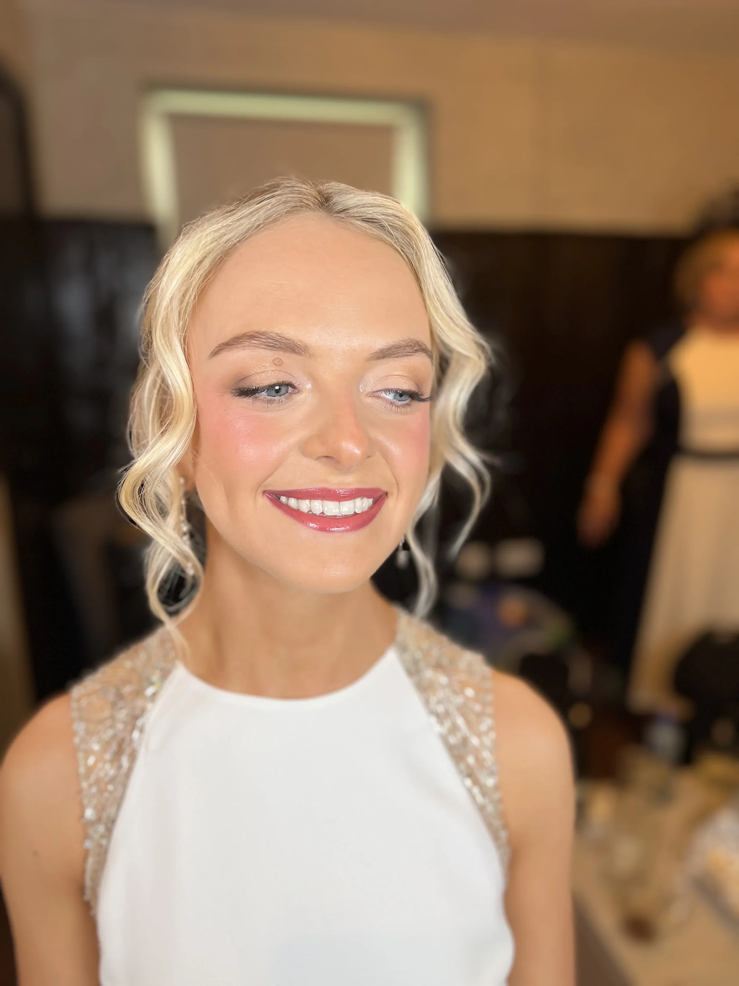 Close-up of a smiling woman and looking down, with light blonde hair, wearing a white wedding dress with beaded embellishments on the shoulders, in an indoor setting.