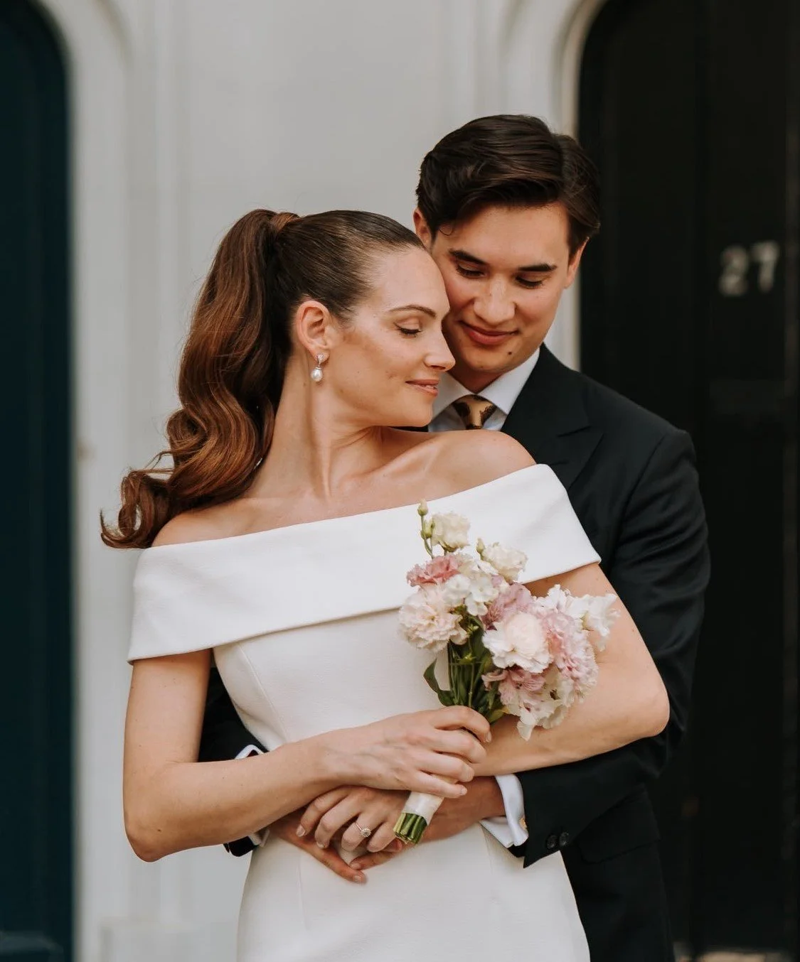 A newly married couple embraces, with the bride holding a bouquet of pink and white flowers, dressed in an off-the-shoulder white wedding gown, and the groom in a black suit with a patterned tie.