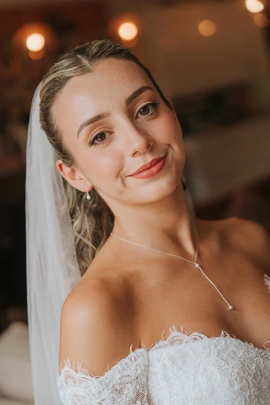 A bride in a white wedding dress with a veil, smiling softly, wearing earrings and a delicate necklace, in an indoor setting with warm lighting. She wears soft glam makeup and hair styled in an elegant half-up style.