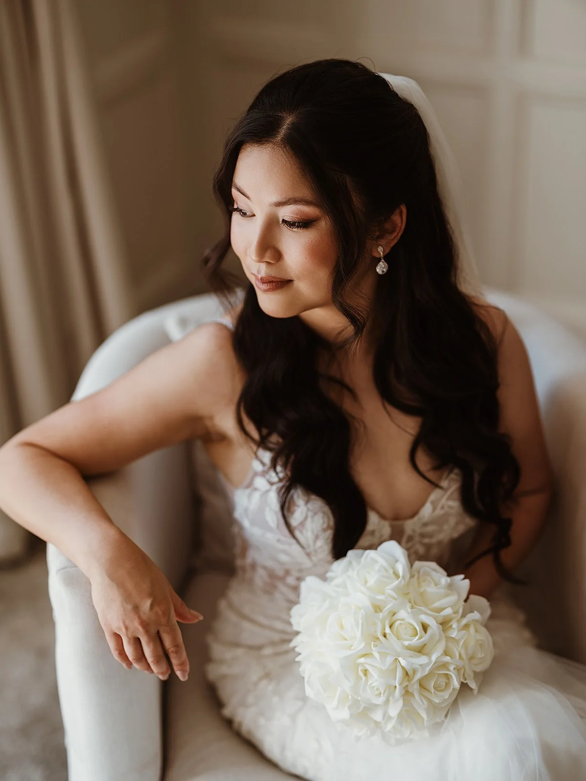 a bride on her wedding day sits in a white armchair, wearing soft glam makeup and romantic, wavy hairstyle