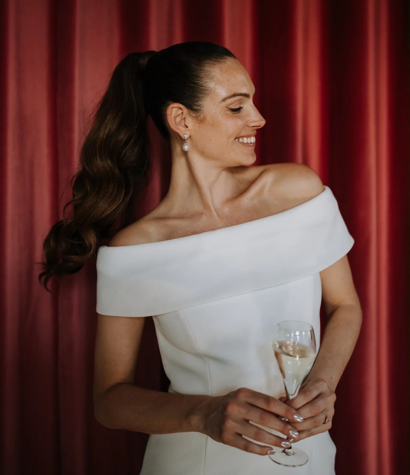 A bride with long dark hair in a ponytail, wearing pearl earrings and a white off-the-shoulder dress, smiling and holding a glass of champagne against a red curtain background.
