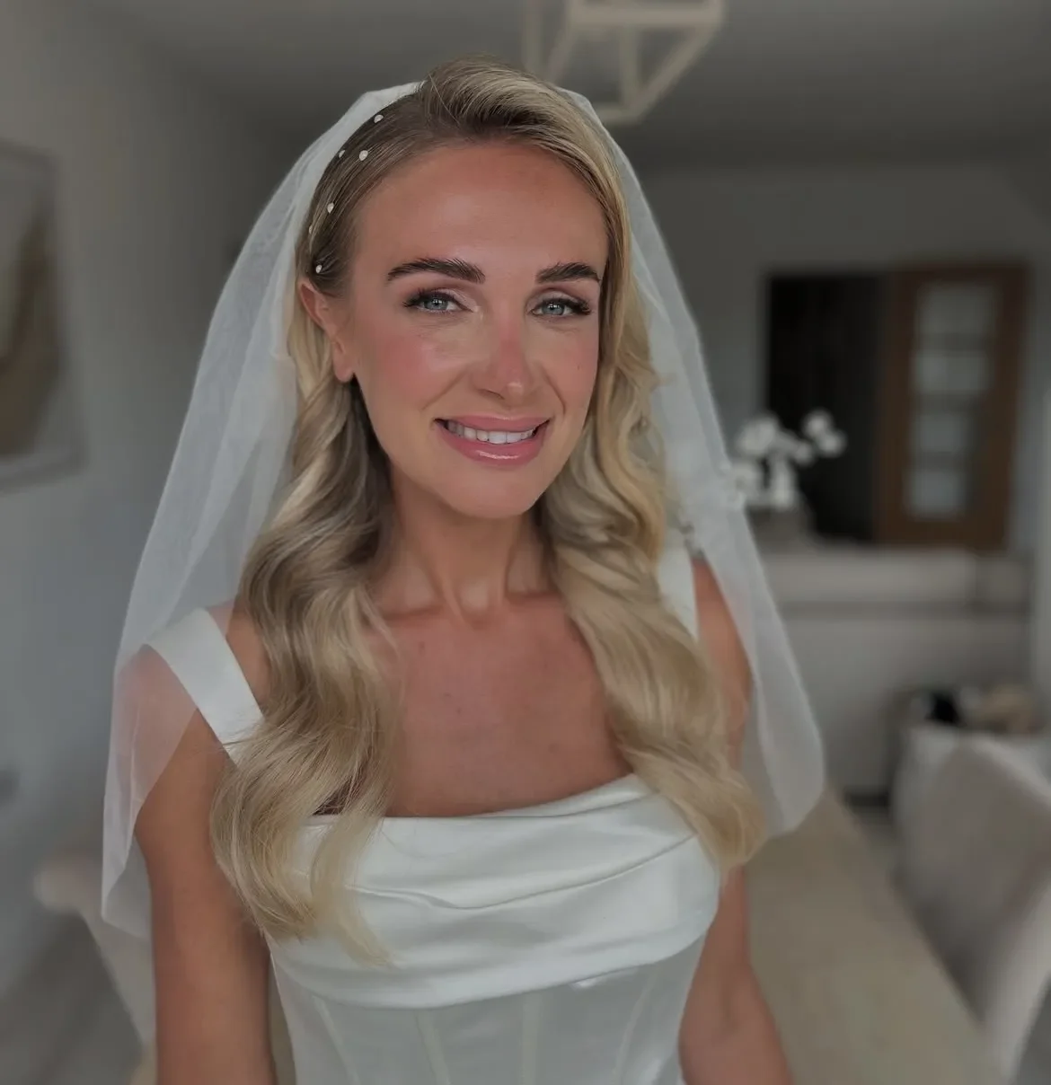 A bride with long blonde hair wearing a white wedding dress and veil, smiling in an indoor setting.