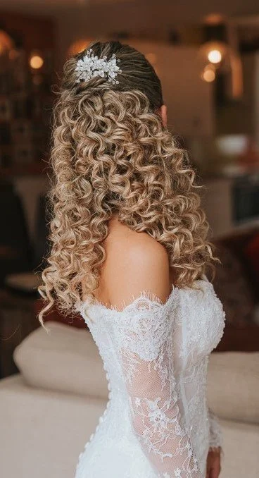 Close-up of a bride with curly, blonde hair wearing a white lace off-the-shoulder wedding dress and a floral hair accessory, indoors.