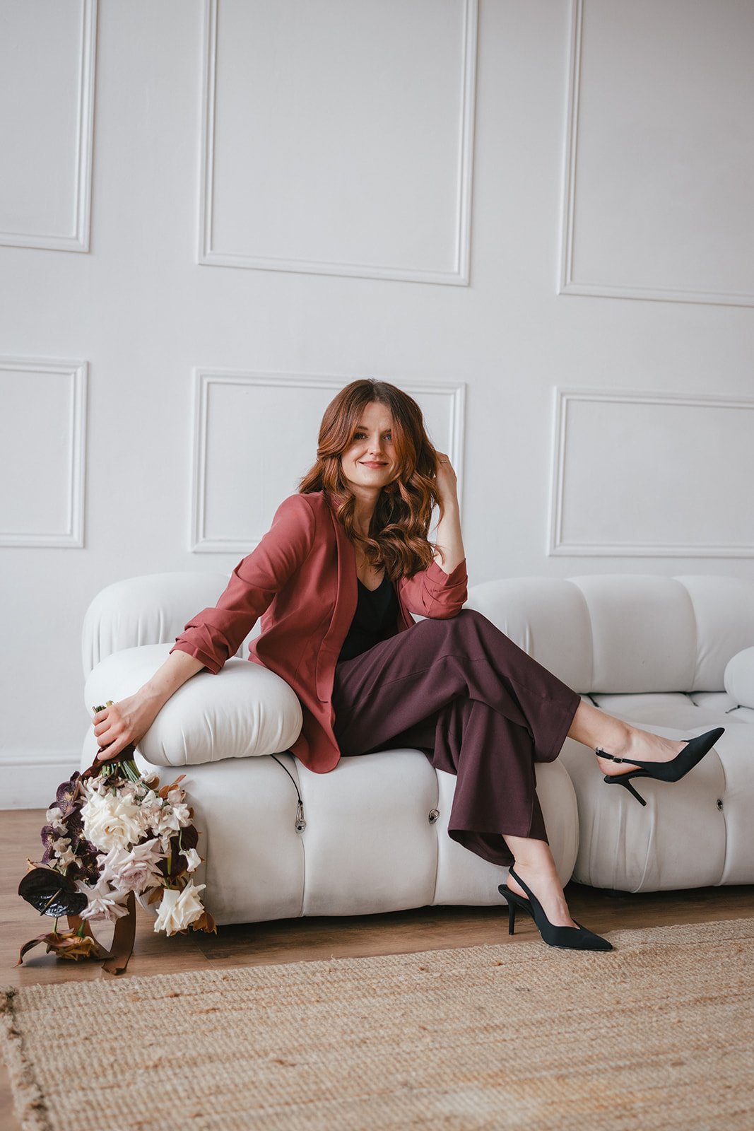 A woman with long wavy red hair sitting on a white sofa, holding a bouquet of flowers, smiling, wearing a rust-coloured blazer, black top, maroon trousers, and black high heels, in a bright room with white wall paneling. For a branding shoot.