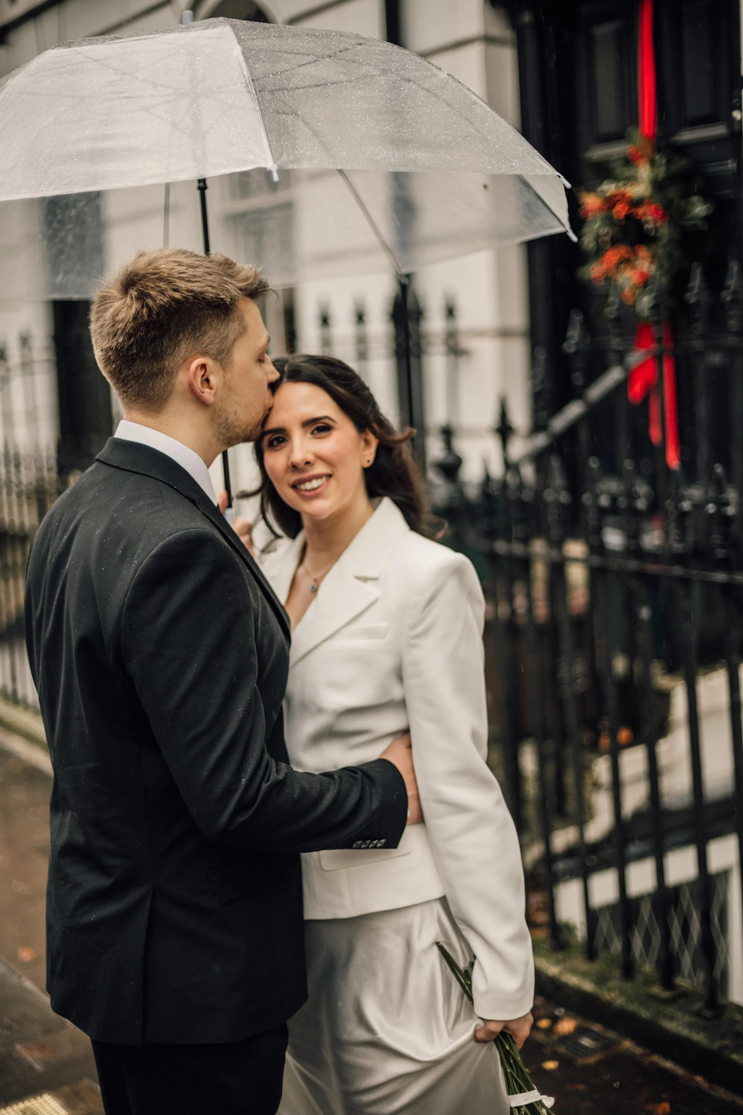 A couple dressed in formal attire embracing under a transparent umbrella in rainy weather, outside a row of black fence and buildings.