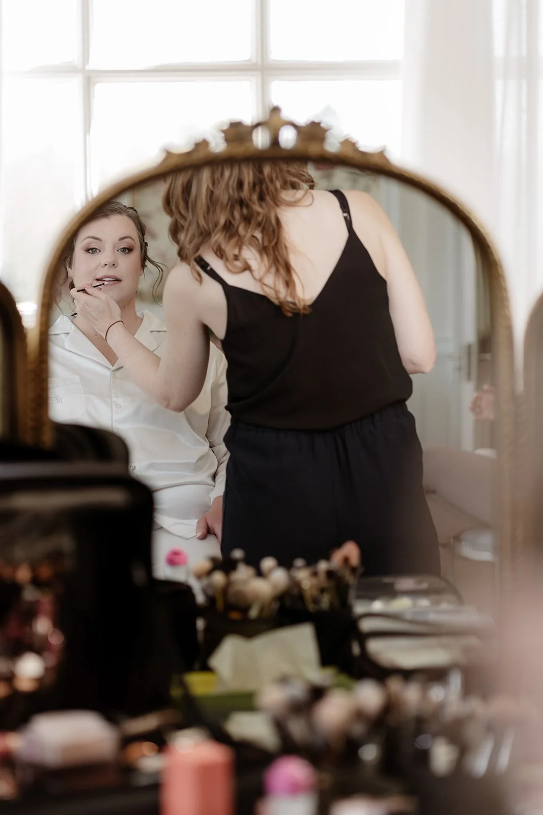 a bride sees herself in a mirror as she has her wedding day lipstick applied as she dits in a makeup chair, wearing white pyjamas