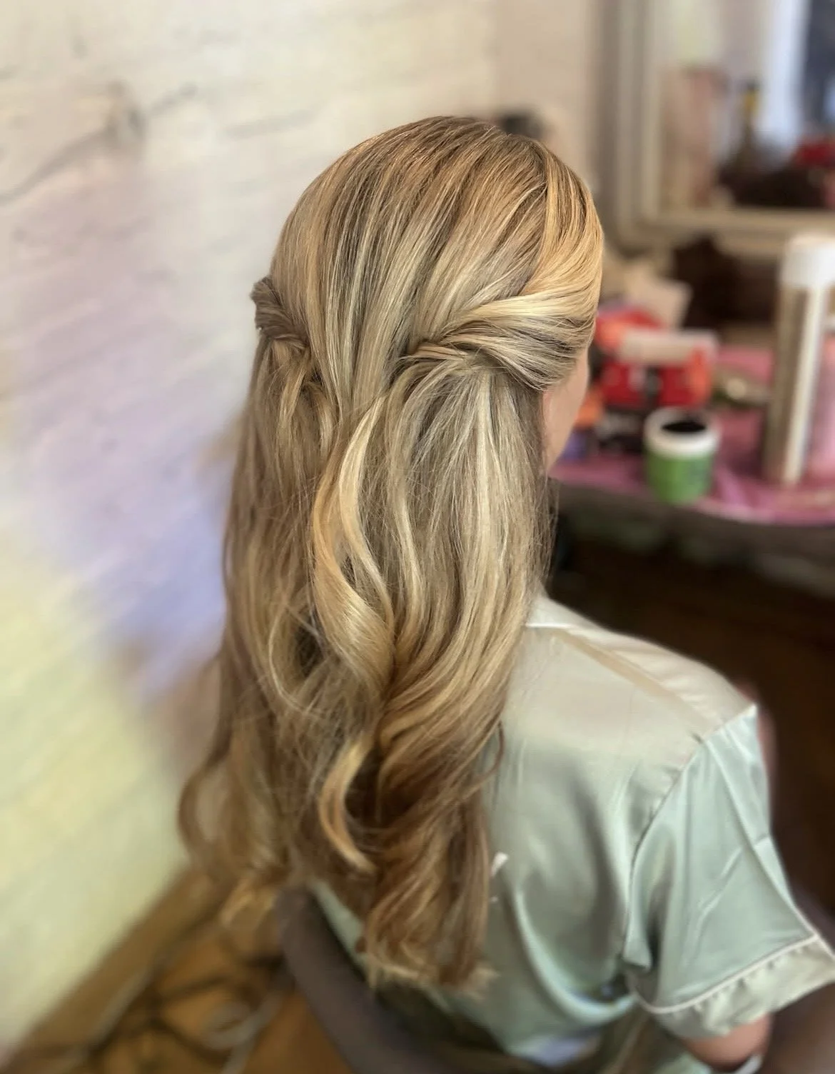 Back view of a woman with long blonde hair styled in loose curls and a twisted half-up hairstyle, sitting in a salon.