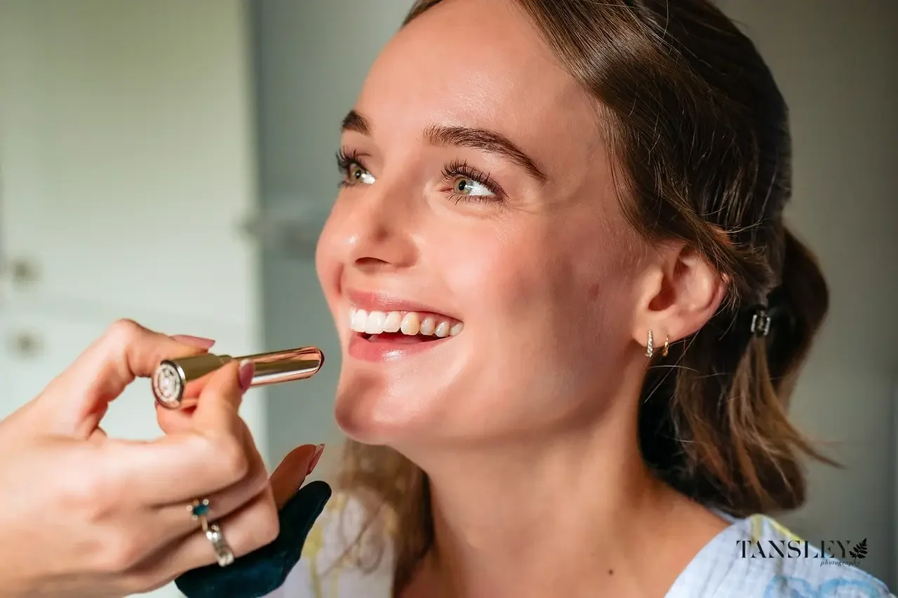 Close-up of a woman smiling as a makeup artist applies lipstick to her lips with a lipstick tube.