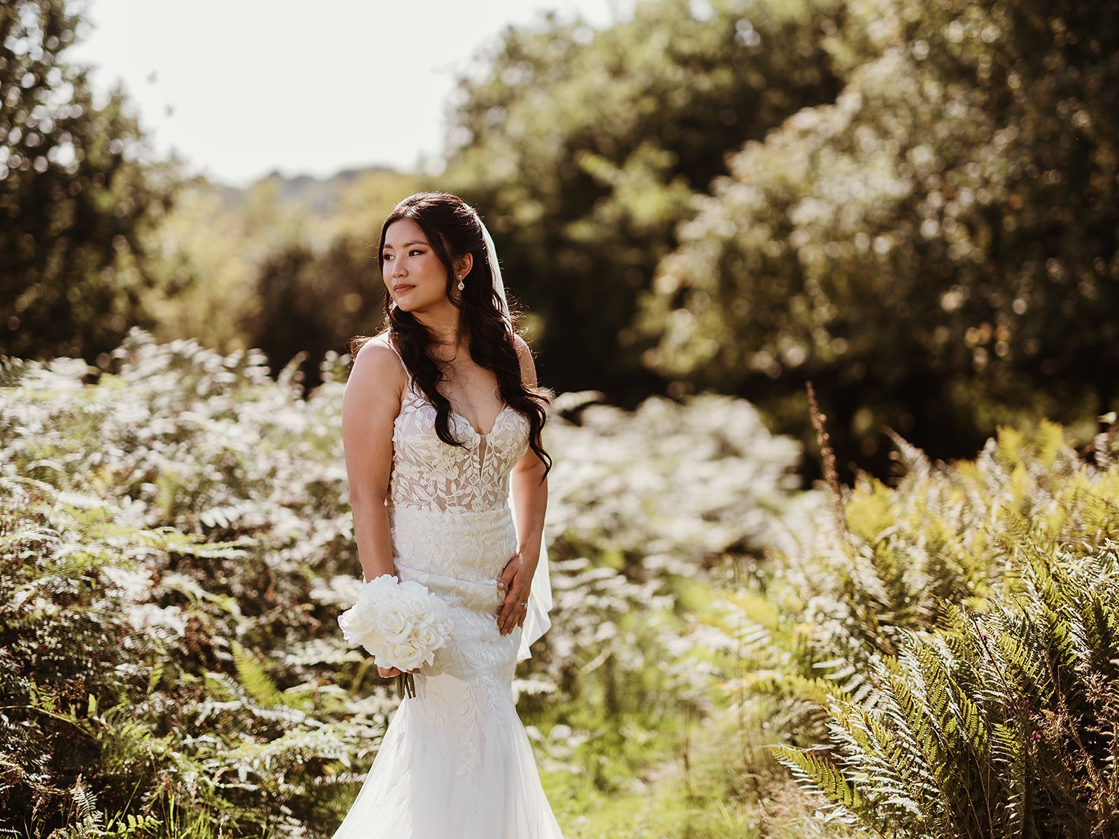 a bride stands against greenery, wearing a white wedding dress and holding her bridal bouquet