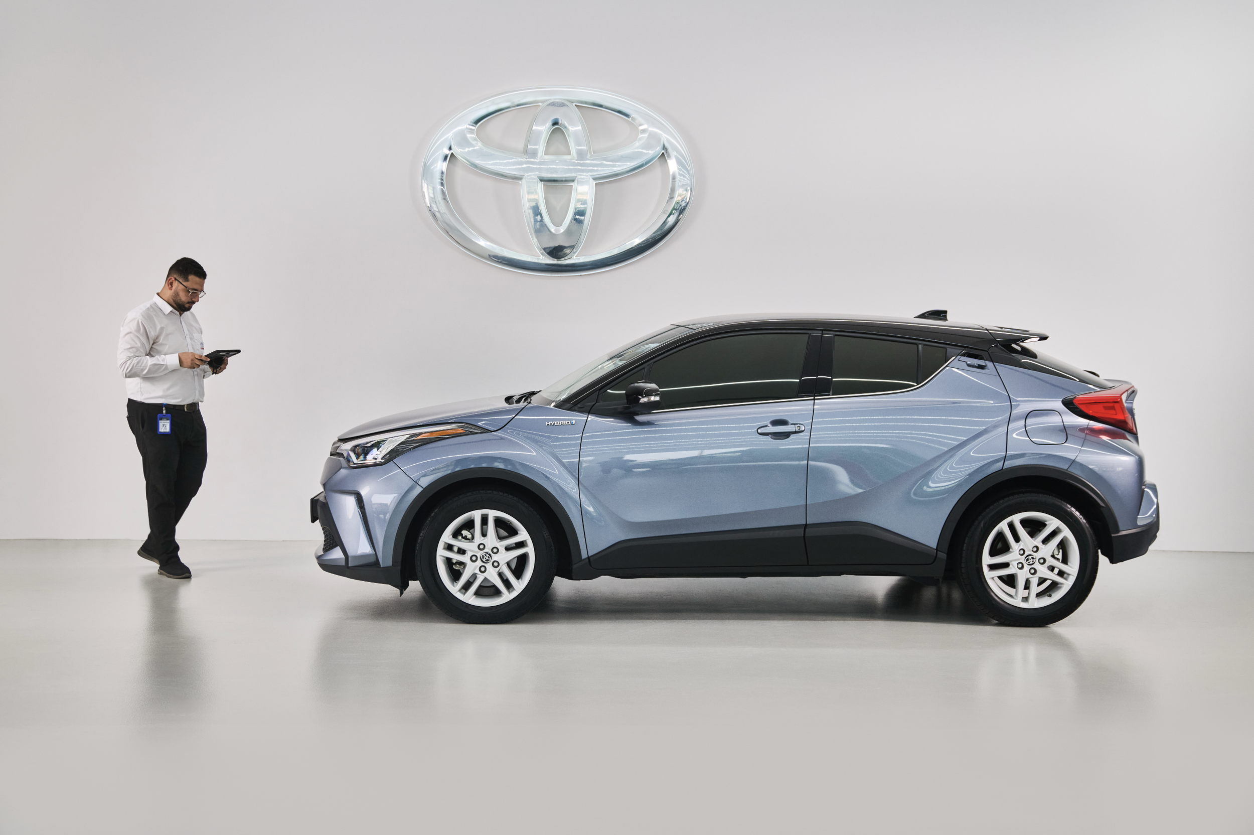A man in white shirt and black pants examining a blue Toyota crossover vehicle inside a showroom with a Toyota logo on the wall.