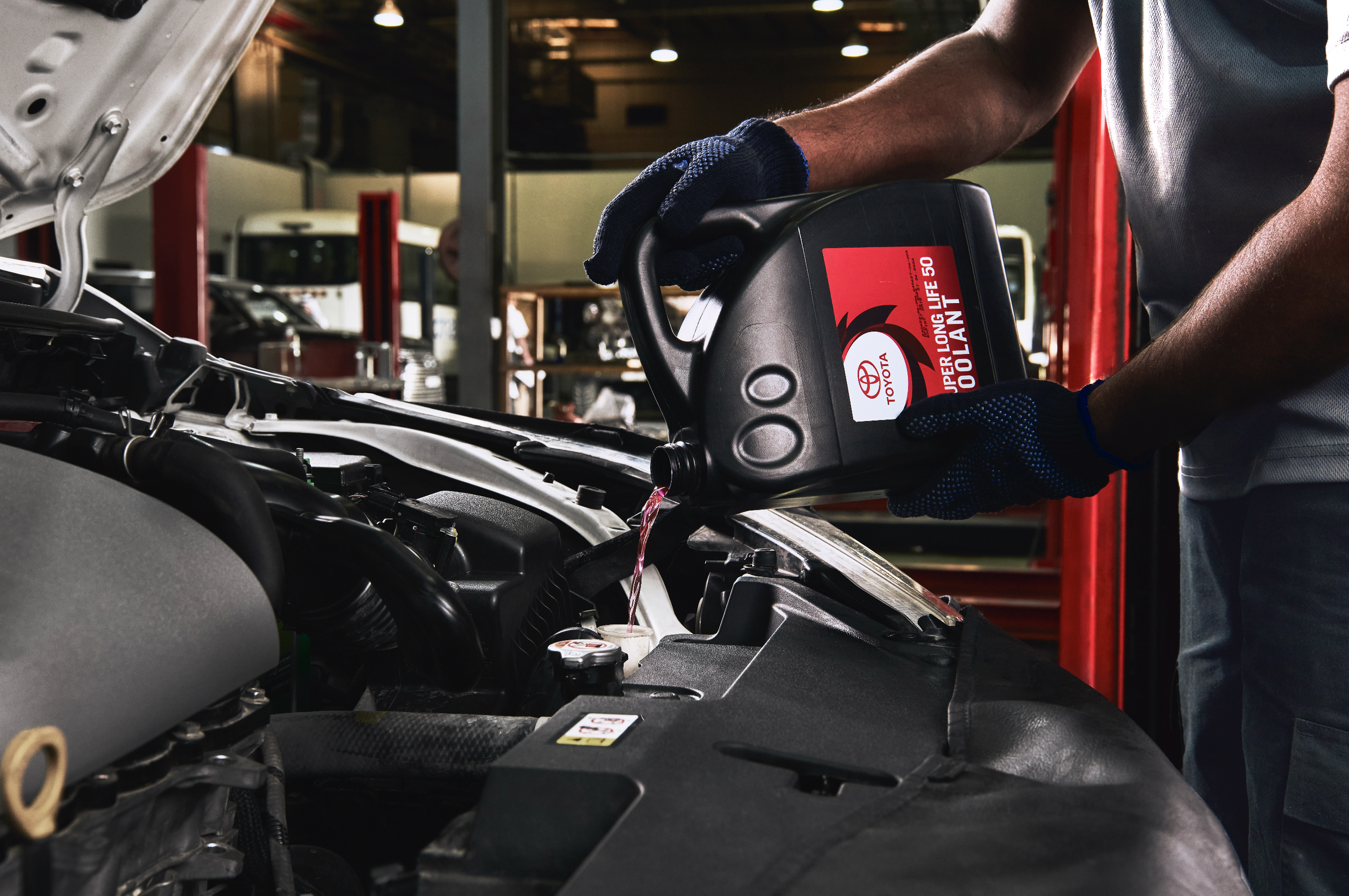 A person is pouring engine oil into a car engine in an auto repair shop. The individual is wearing dark gloves and a gray shirt, and the background shows other vehicles and workshop equipment.