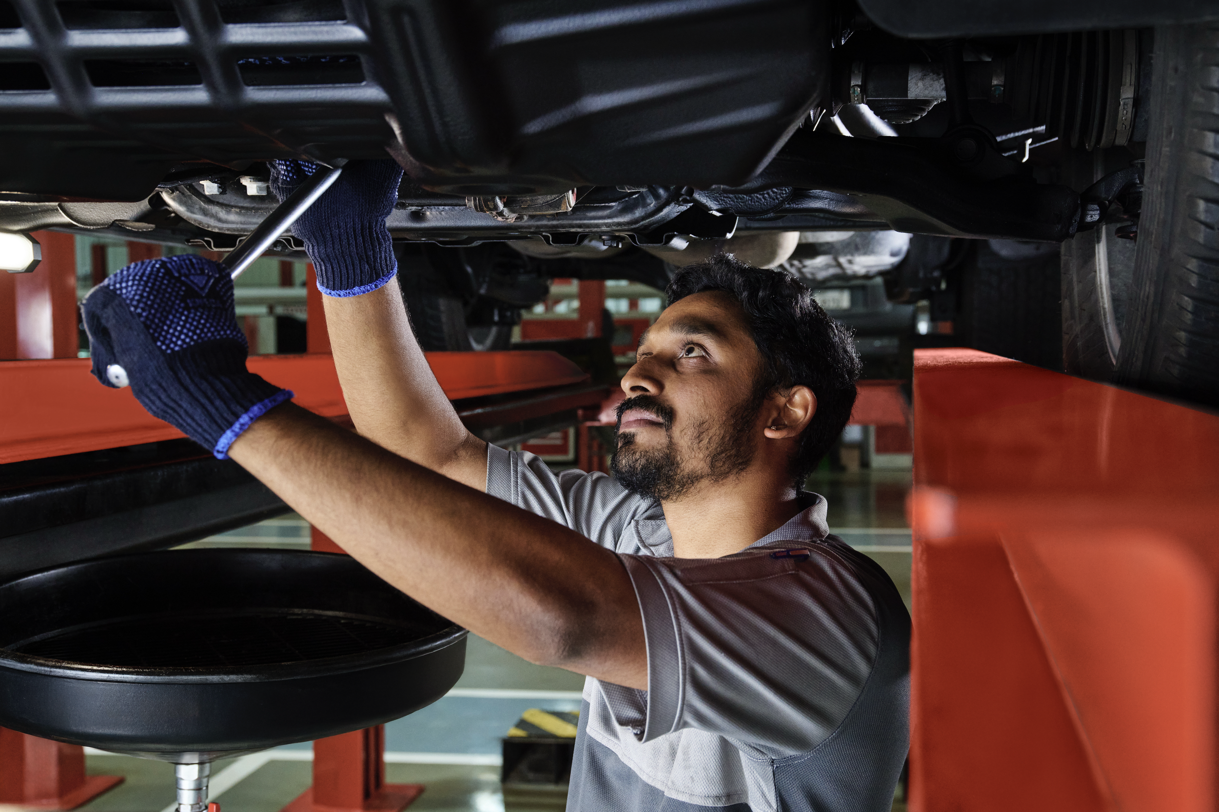 A mechanic working underneath a vehicle on a hydraulic lift, wearing gloves and inspecting or repairing the underside of the car