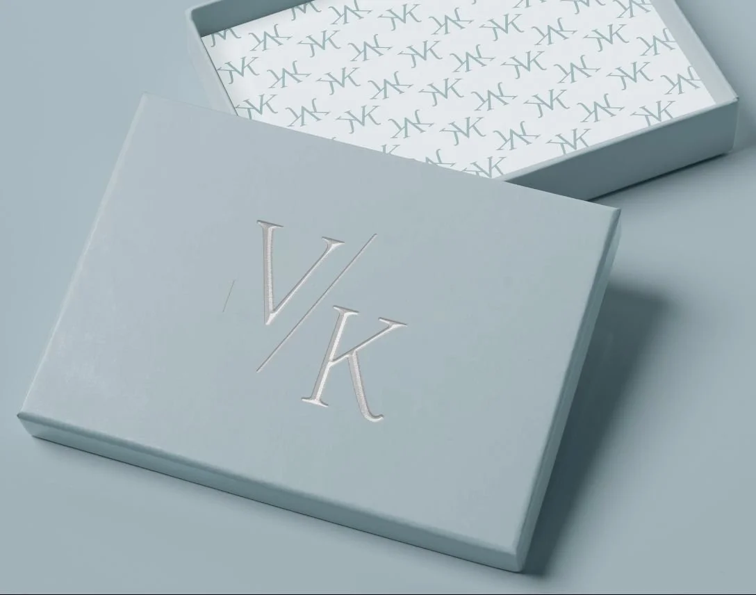 A close-up of a pale blue box with the letters 'V and K' engraved on its side. The inside of the box is visible, revealing a repeating pattern of interlinked "VK" motifs.