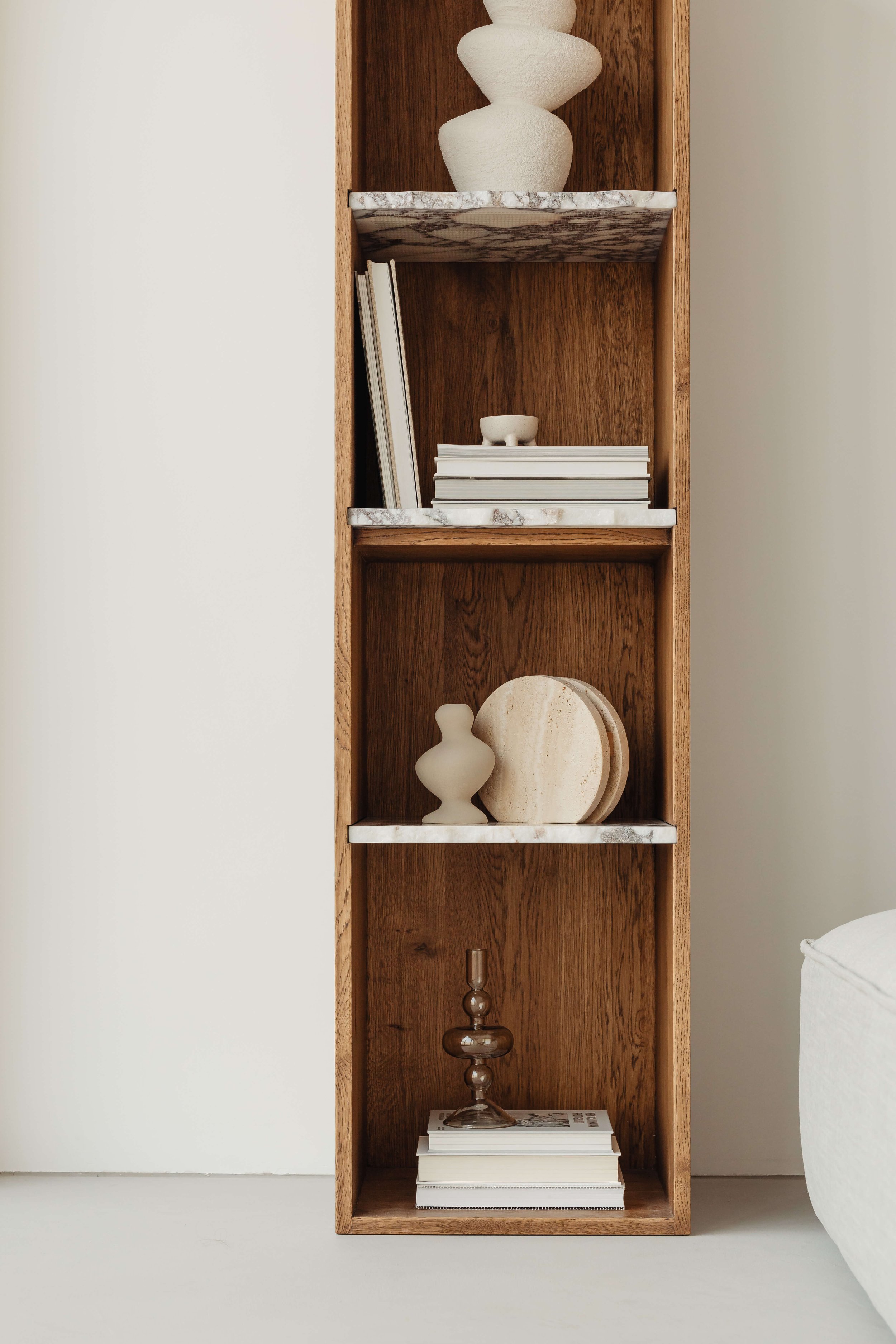 A tall, narrow wooden bookshelf stands against a white wall. The bookshelf has four shelves with marble-patterned tops. The top shelf holds a textured white sculpture, followed by a book and a small bowl on the second shelf.