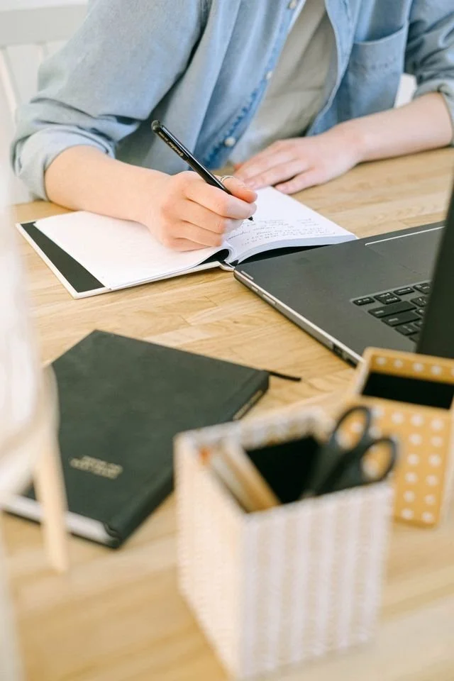 Person journaling at a wooden desk with a laptop and stationery in a bright studio.
