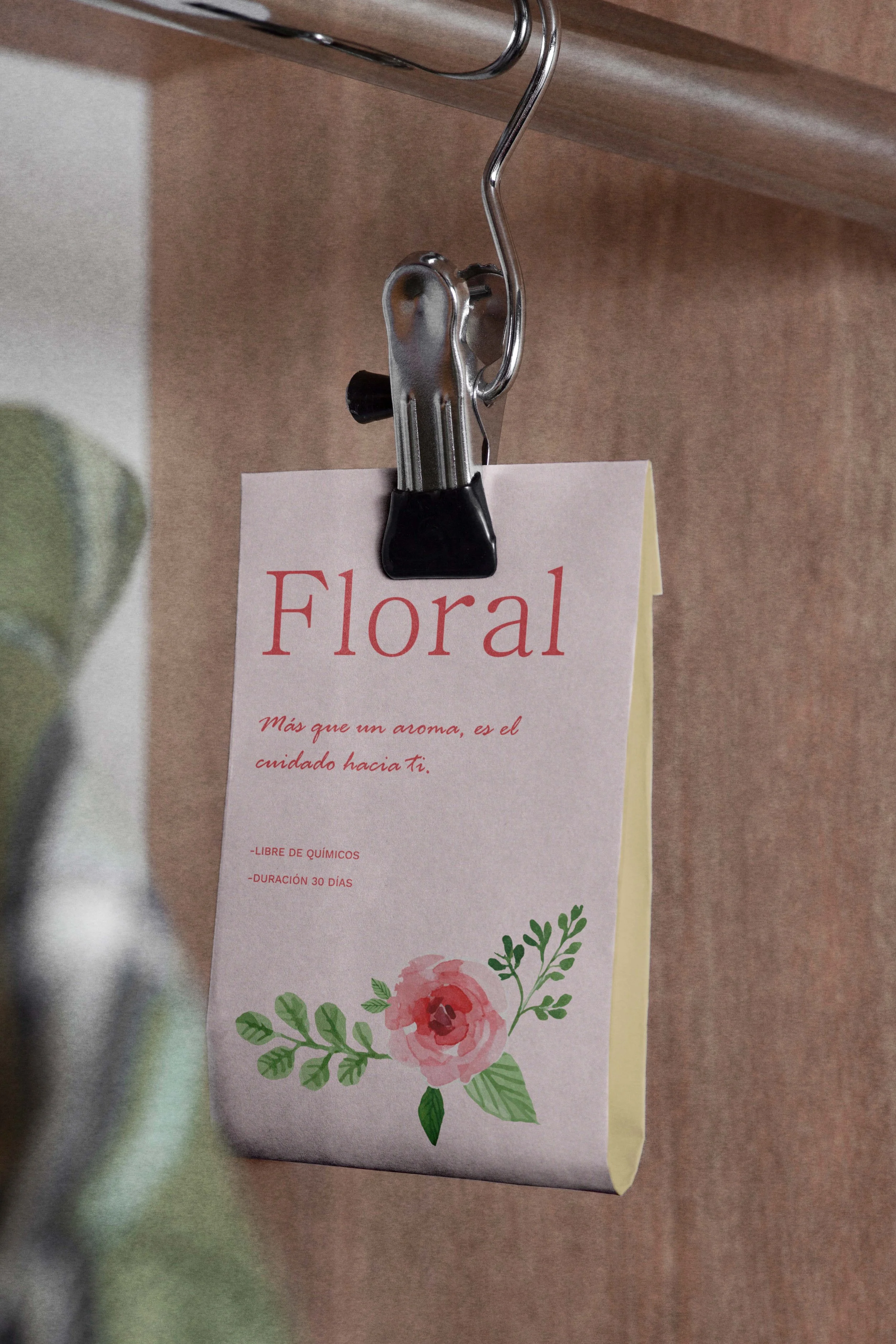 A minimalist hanging air freshener for the boutique, featuring a delicate floral watercolour illustration and 'Floral' typography, suspended by a silver metal clip against a natural wood background.