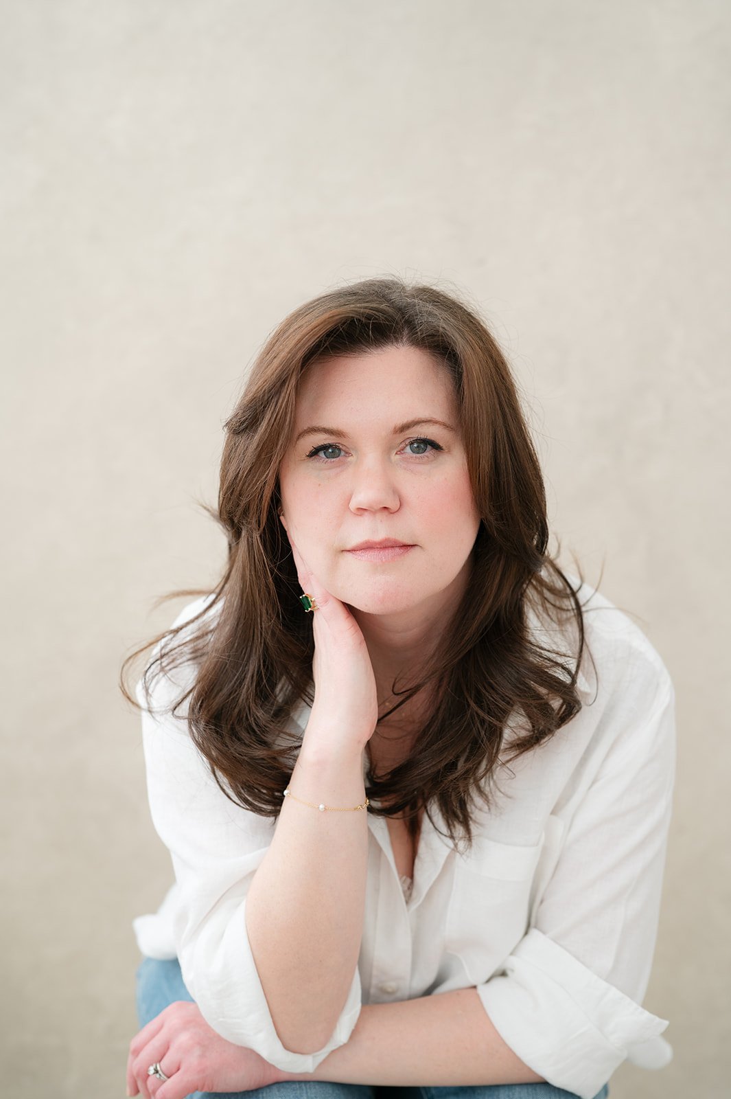 A woman with shoulder-length brown hair and blue eyes, wearing a white shirt, sitting with her chin resting on her hand, against a light-colored textured background.