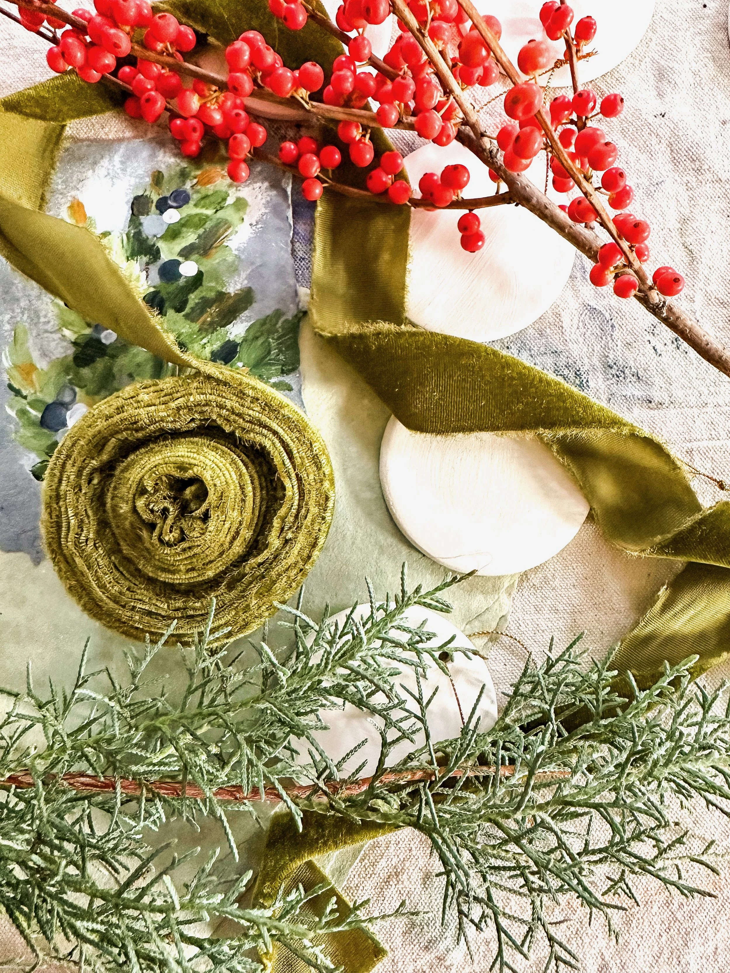 Decorative arrangement with red berries, white ceramic spheres, green fabric ribbons, and green foliage on a beige cloth background.