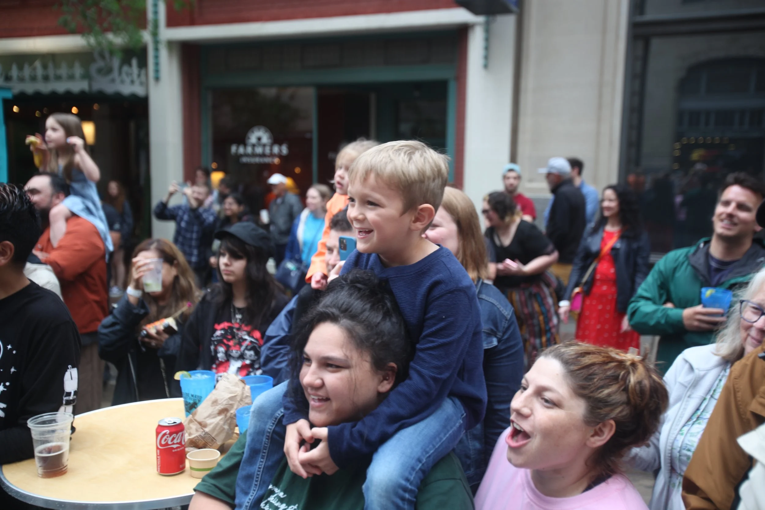 Young boy on a woman's shoulders smiling during a crowded outdoor gathering