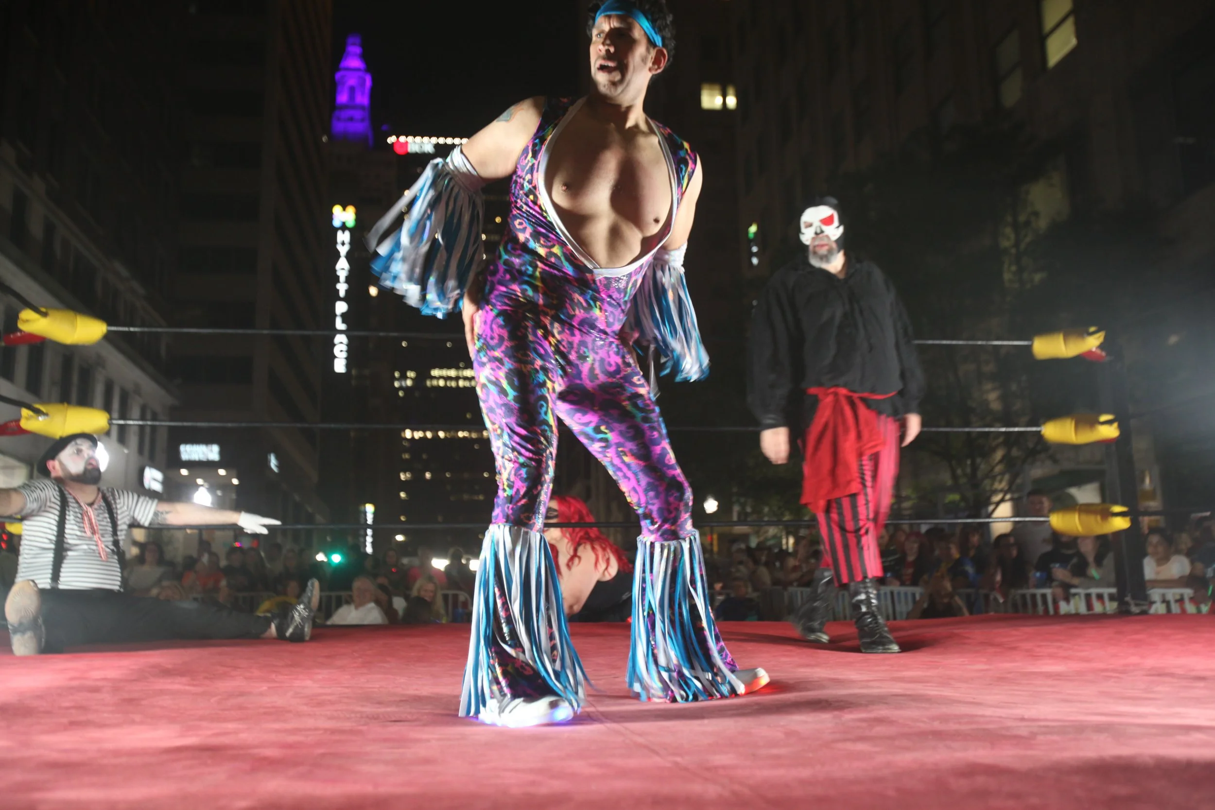 Professional wrestling match taking place outside at night in an urban setting with tall buildings and city lights. One wrestler is dressed in colorful, patterned disco-style attire with matching bell-bottoms, and is in the center of the ring. Two ot