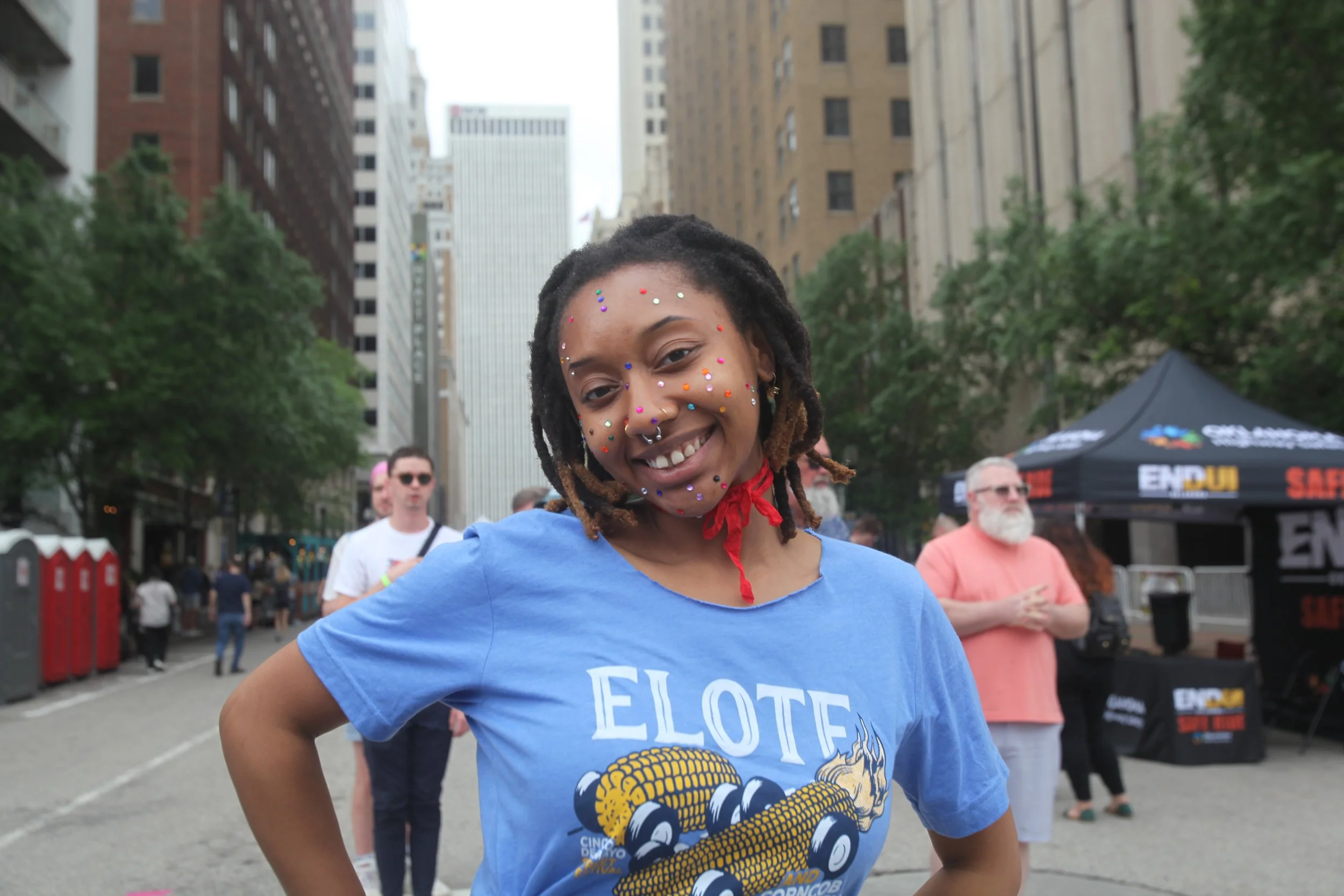Smiling young Black woman with dreadlocks, decorated with colorful face gems, wearing a blue t-shirt that says 'ELOTE' and features a corn graphic, at an outdoor event on a city street with tall buildings in the background and people gathered around.