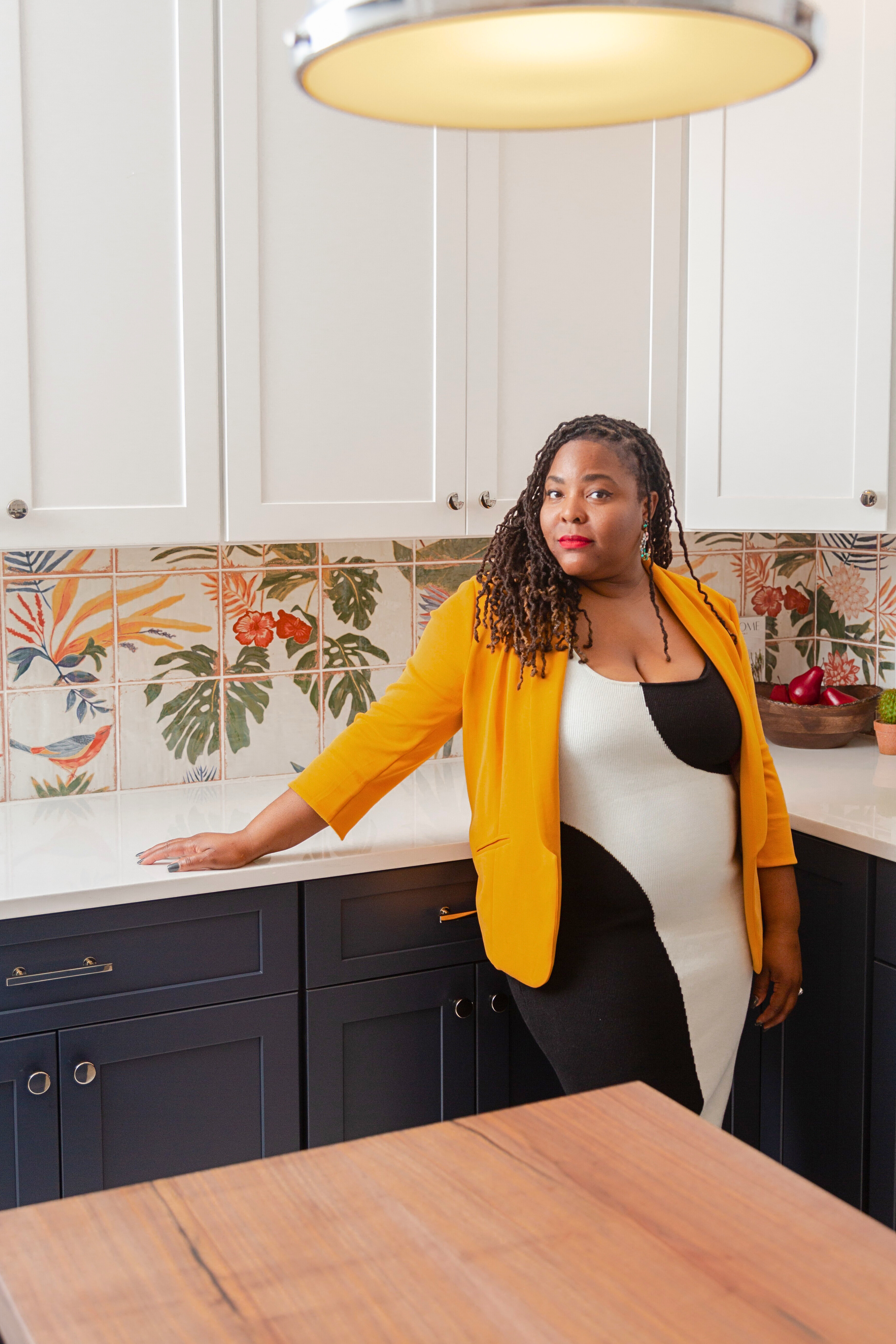 A woman in a black and white dress with a yellow blazer standing in a kitchen, resting her hand on the kitchen counter, with floral backsplash and dark cabinets.