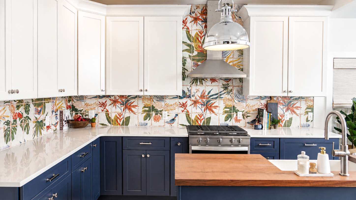 Modern kitchen with white upper cabinets, dark blue lower cabinets, tropical patterned tile backsplash, stainless steel stove, wooden island, and stainless steel range hood.