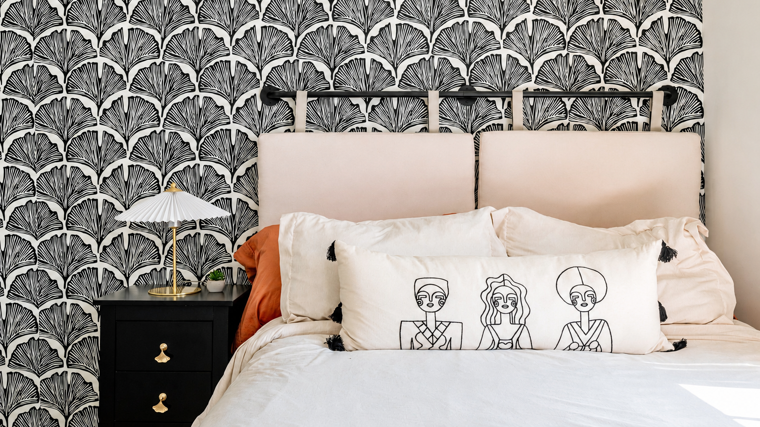 A bedroom with black and white wallpaper featuring a fan pattern, a black nightstand with a small plant, a white lamp, cream-colored headboard, and decorative pillows including one with black line art of three women.