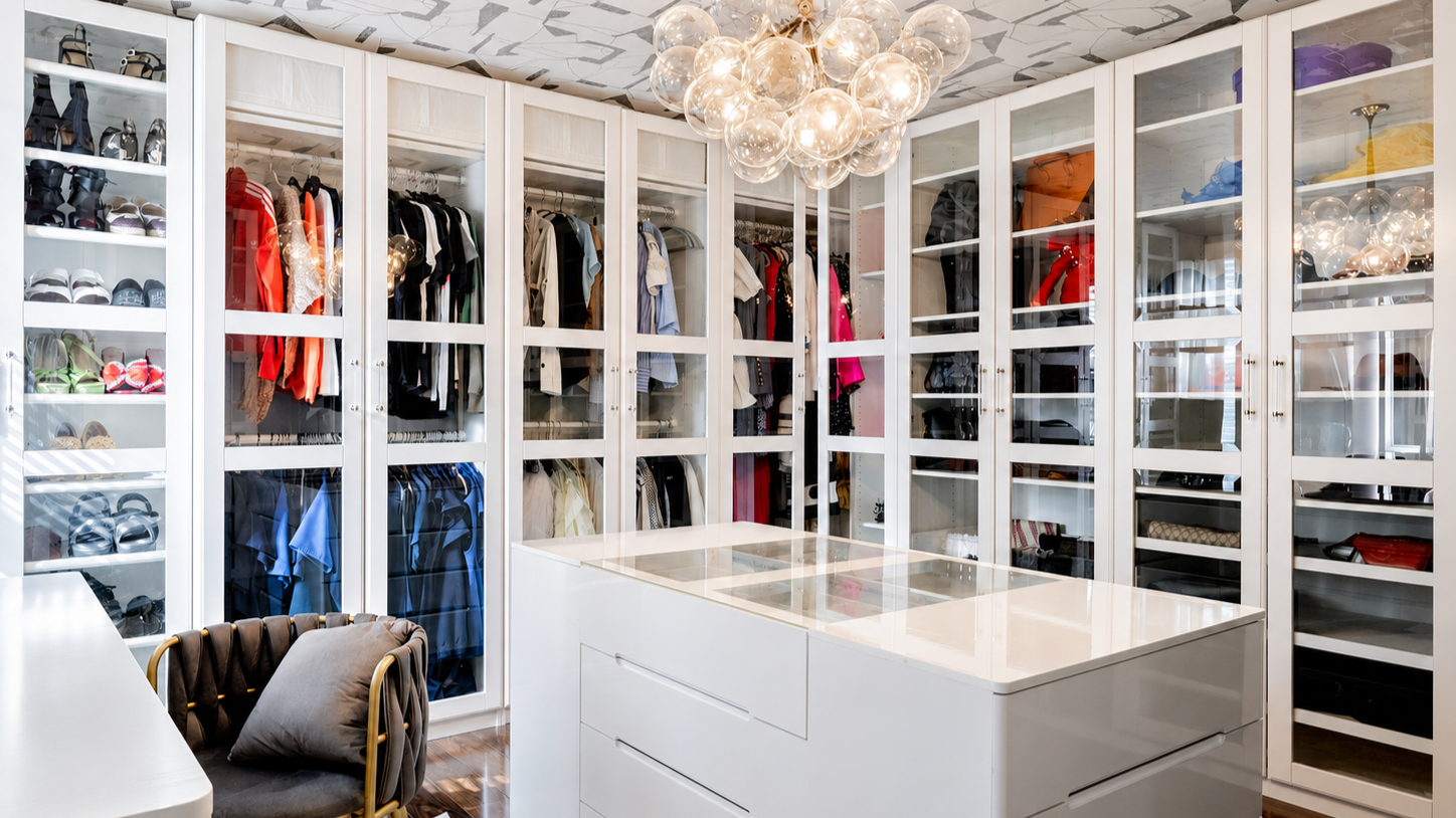 A modern walk-in closet with white cabinetry, clothing and shoes organized behind glass doors, a white island with storage drawers, a chair with a cushion, and a contemporary chandelier with glass globes hanging from the ceiling.