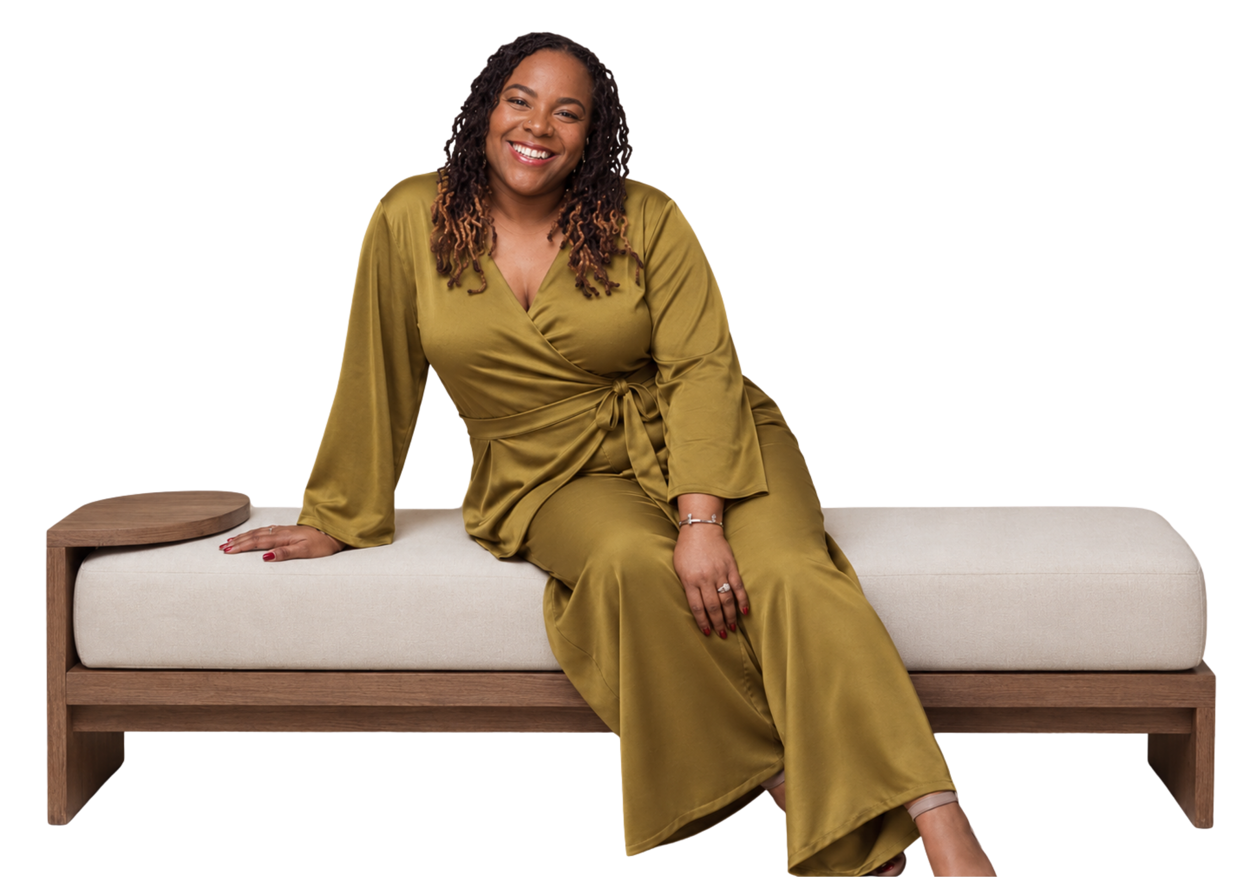 A woman smiling and sitting on a sofa, wearing a gold satin wrap dress, with her hand resting on the sofa and her legs crossed at the ankle.
