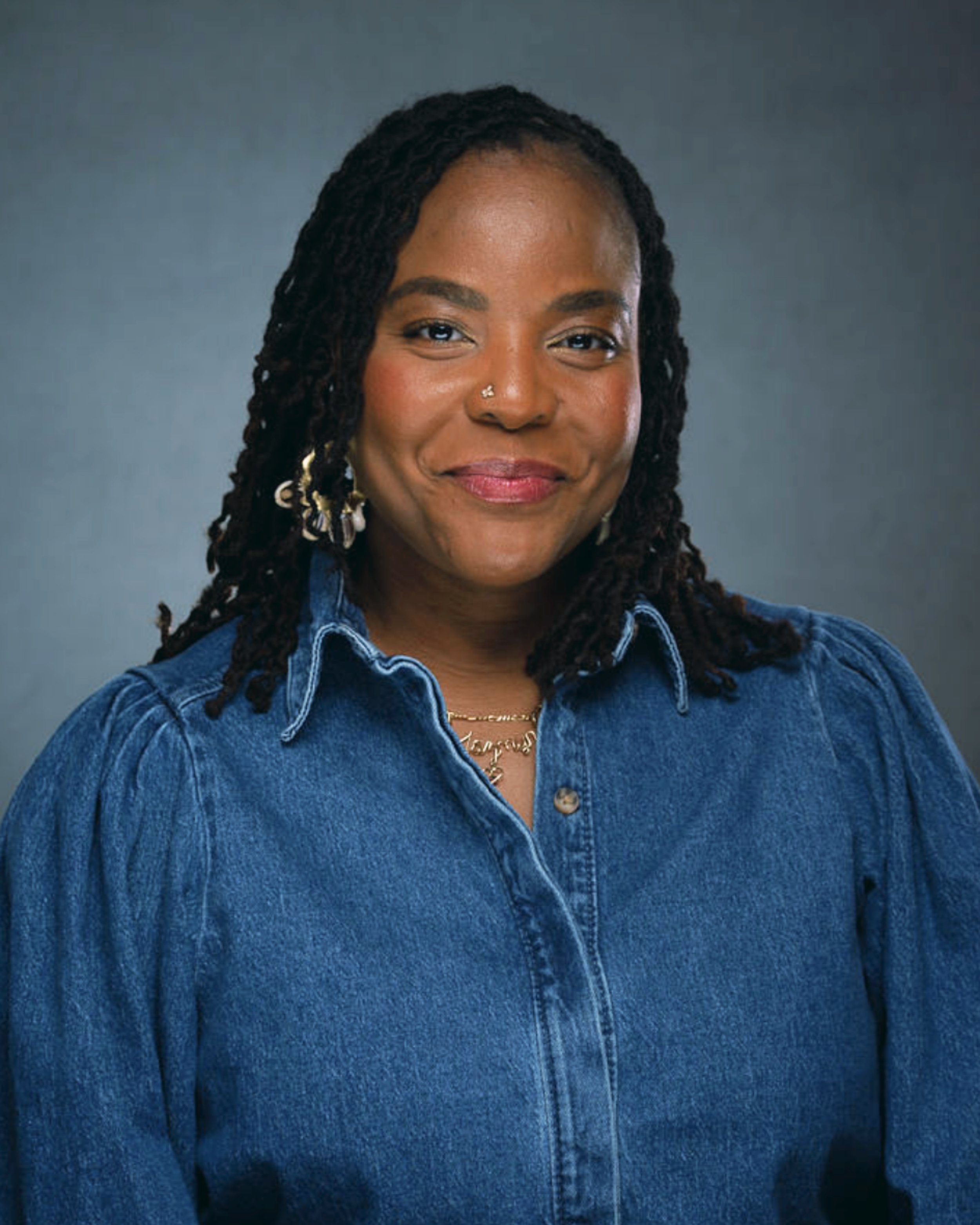 Close-up of a smiling woman with dark curly hair, wearing a denim shirt and earrings, against a plain background.
