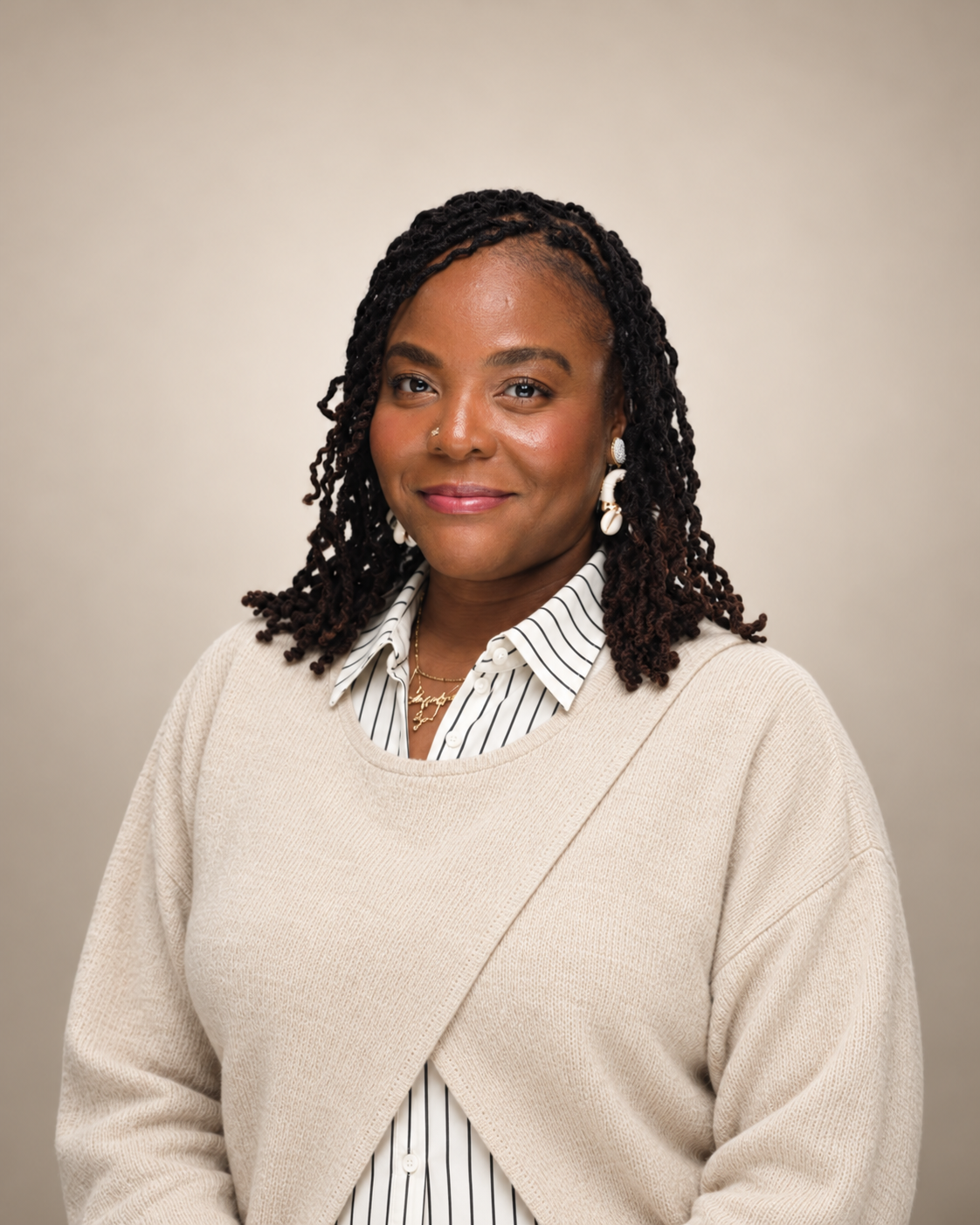 A woman with dark curly hair, wearing a beige sweater over a striped collared shirt, gold jewelry, and earrings, smiling at the camera against a plain background.