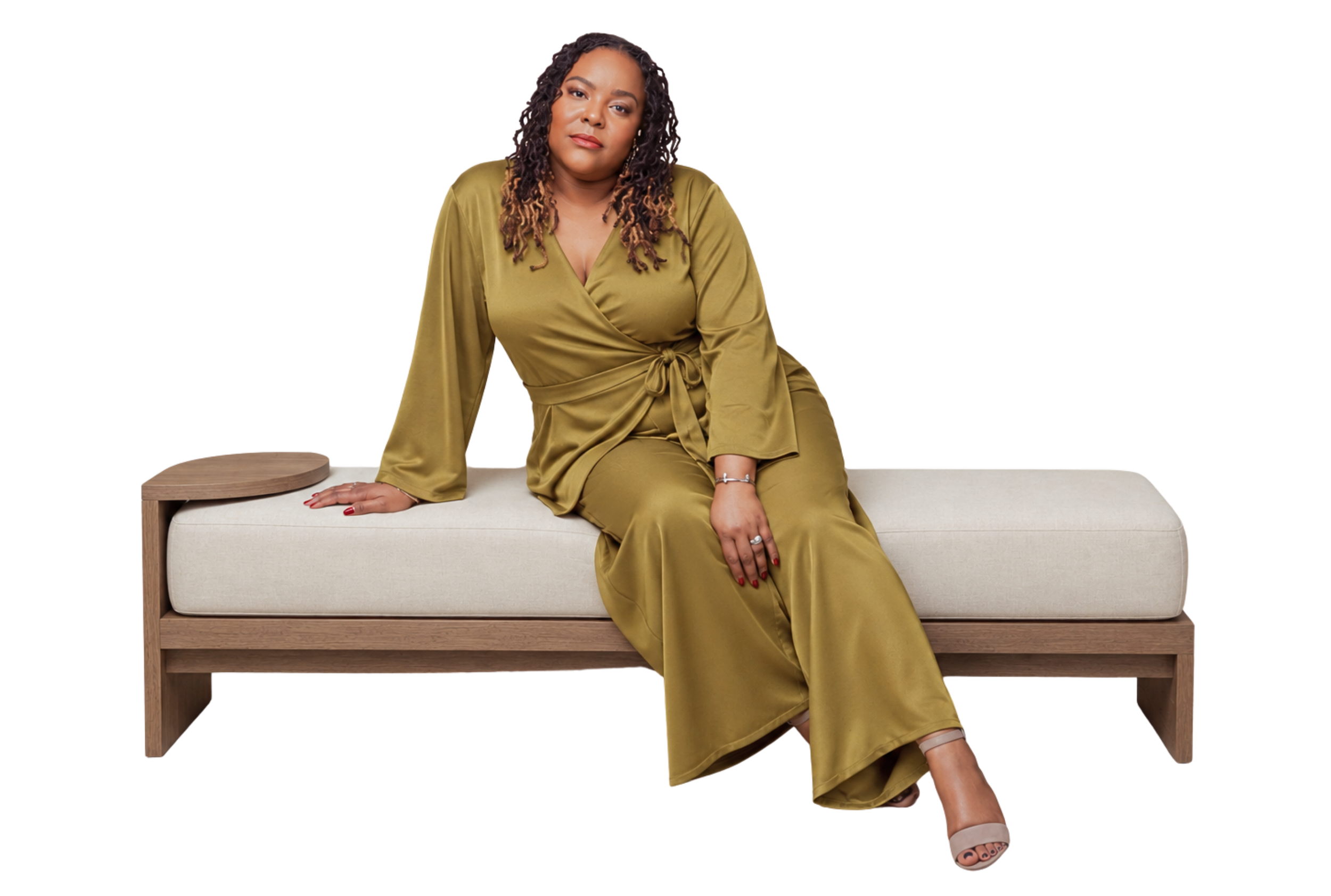 A woman with curly hair wearing a gold satin dress sitting on a modern beige and wood bench, with one hand resting on the seat and the other on her lap, looking at the camera with a neutral expression.