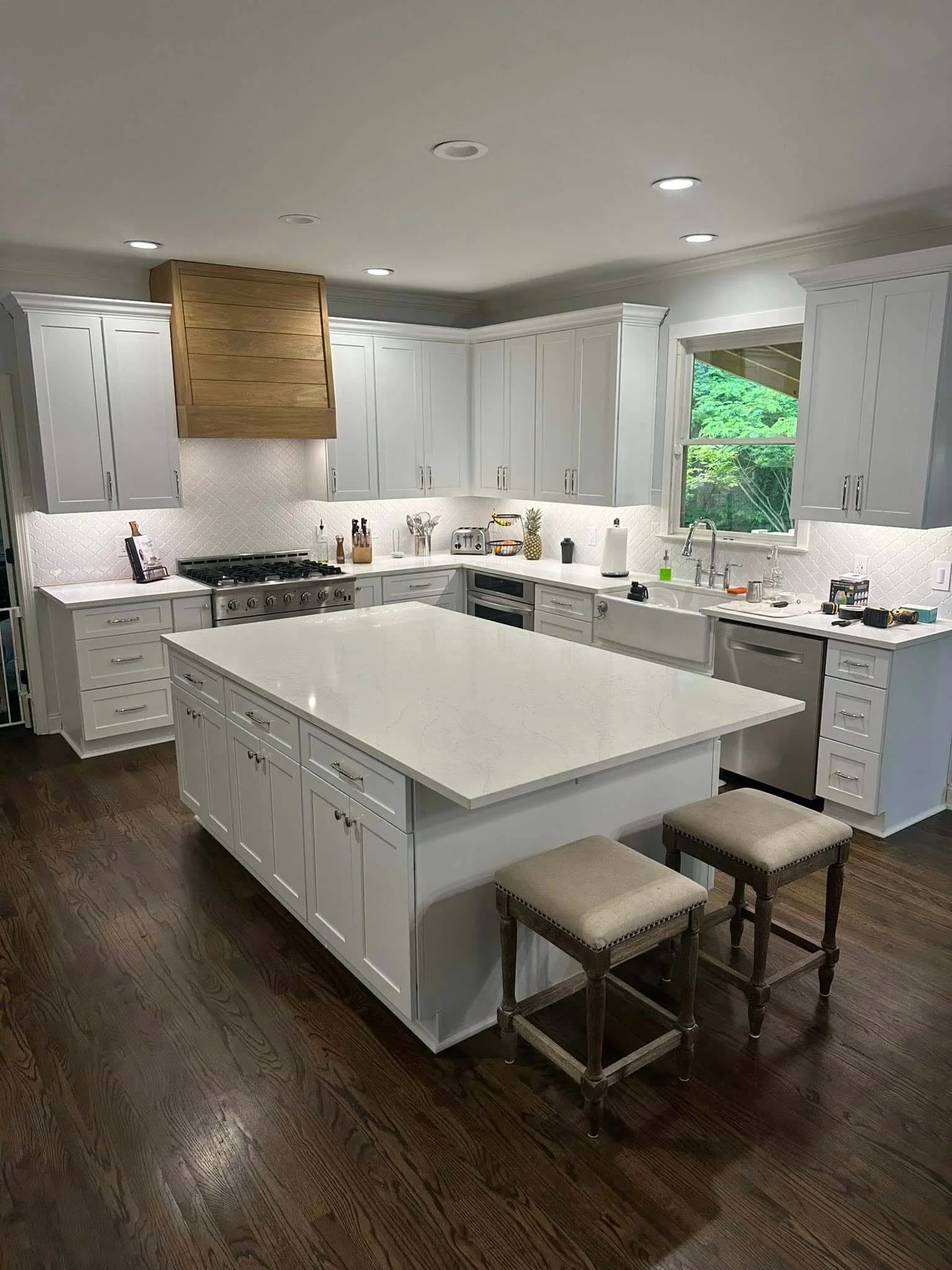 A bright modern kitchen with white cabinets, a large white kitchen island, dark hardwood floors, and a window over the sink with outdoor greenery visible. Two beige stools are in front of the island.