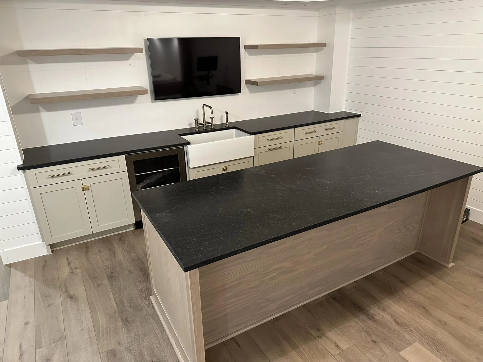 Kitchen with black countertops, white cabinets, a farmhouse sink, a wall-mounted TV, open shelves, and a kitchen island with a black countertop.