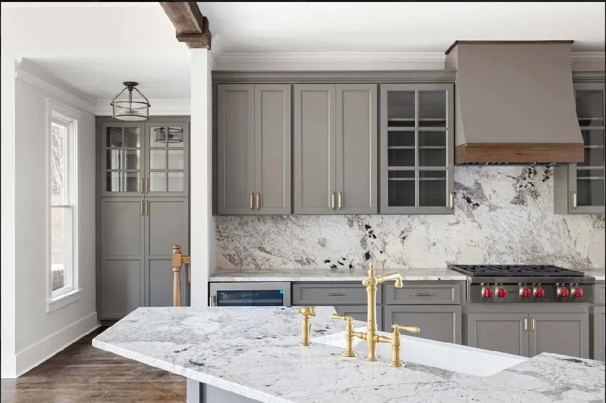 Modern kitchen with gray cabinets, white and gray marble backsplash, gold fixtures, and a large marble kitchen island with a double sink.