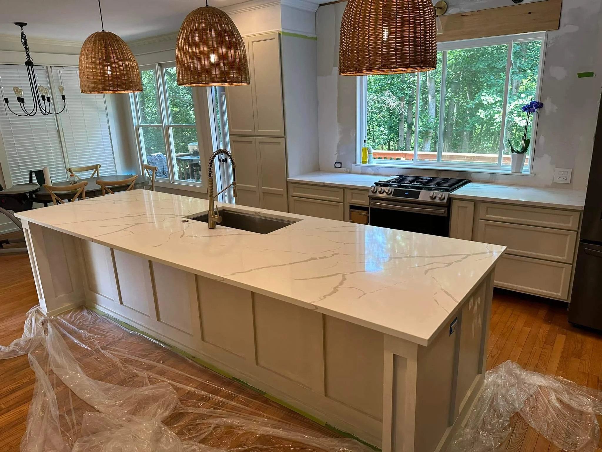 Kitchen with a large white marble island, beige cabinets, a stove at the window, and pendant lights over the island. There is plastic on the floor, indicating renovation.