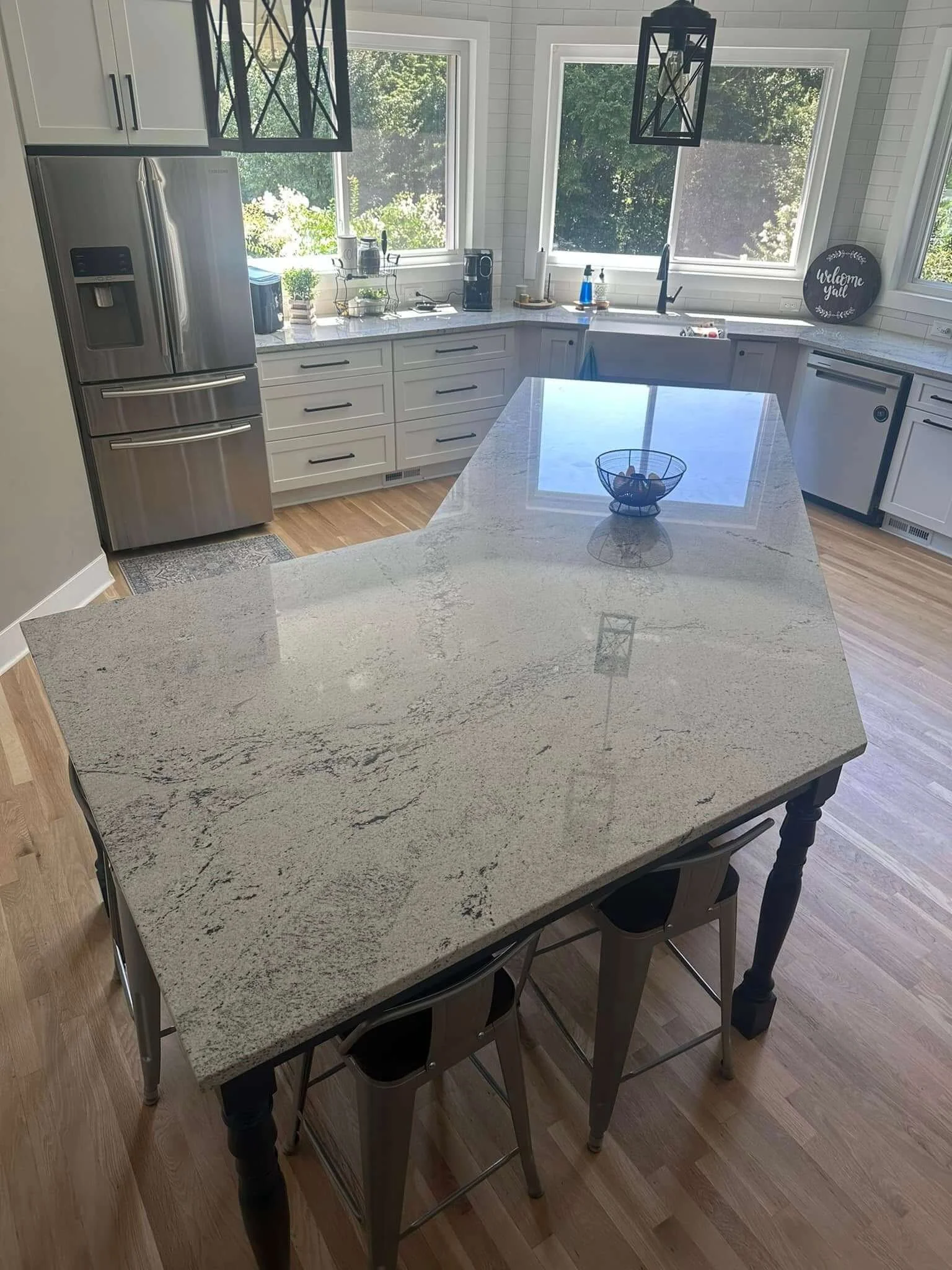 Modern white kitchen with large granite countertop island, stainless steel refrigerator, white cabinetry, and large windows with greenery outside.