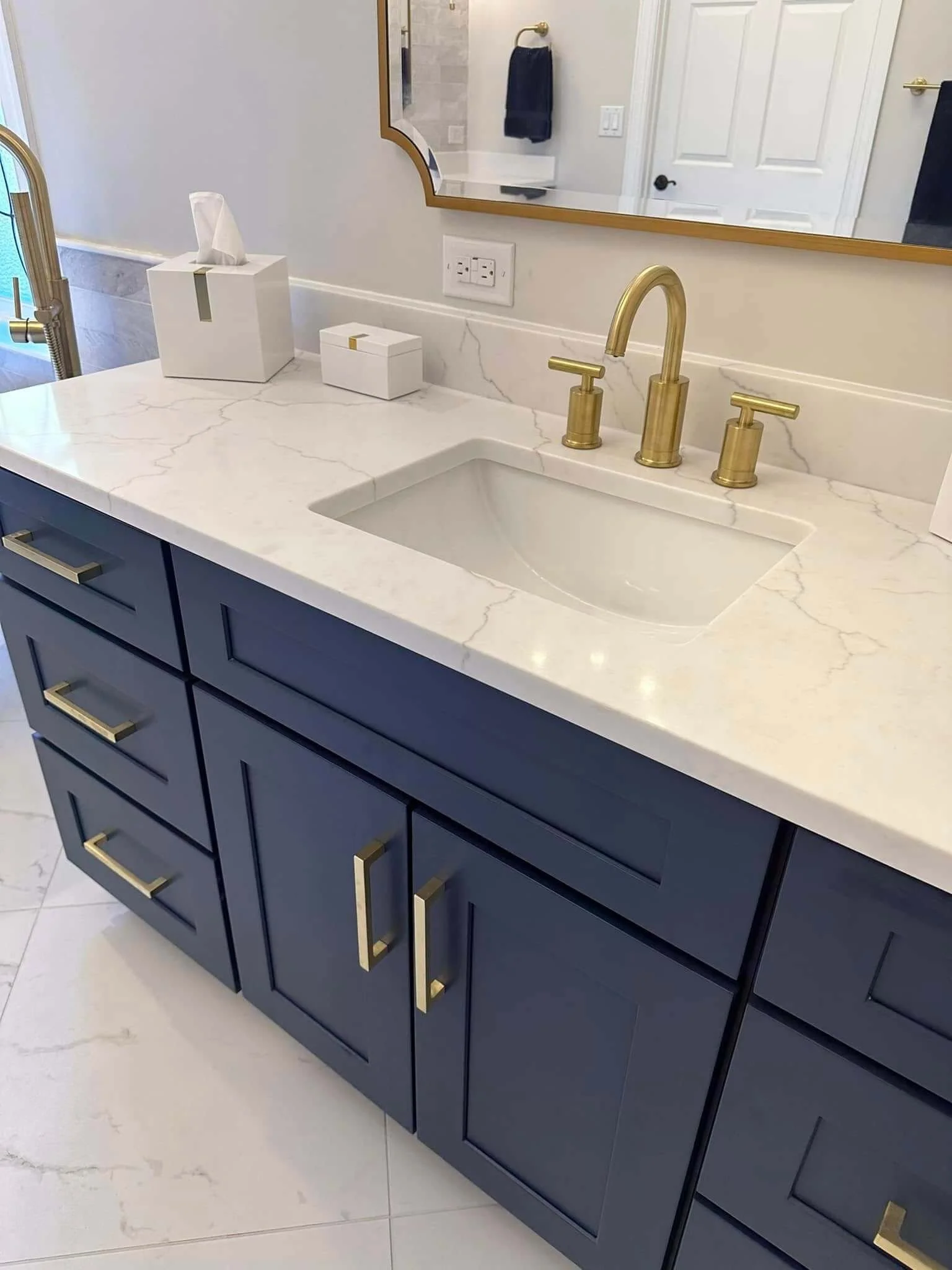 Bathroom vanity with a white quartz countertop, blue cabinetry with gold handles, and a gold faucet. There are tissue boxes and a mirror above the sink.