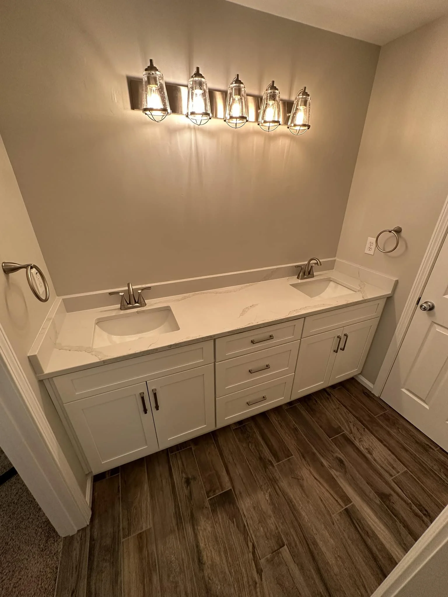 Bathroom vanity with a marble countertop, double sinks, and silver faucets, with a wall-mounted light fixture above, and a mirror absent from view.