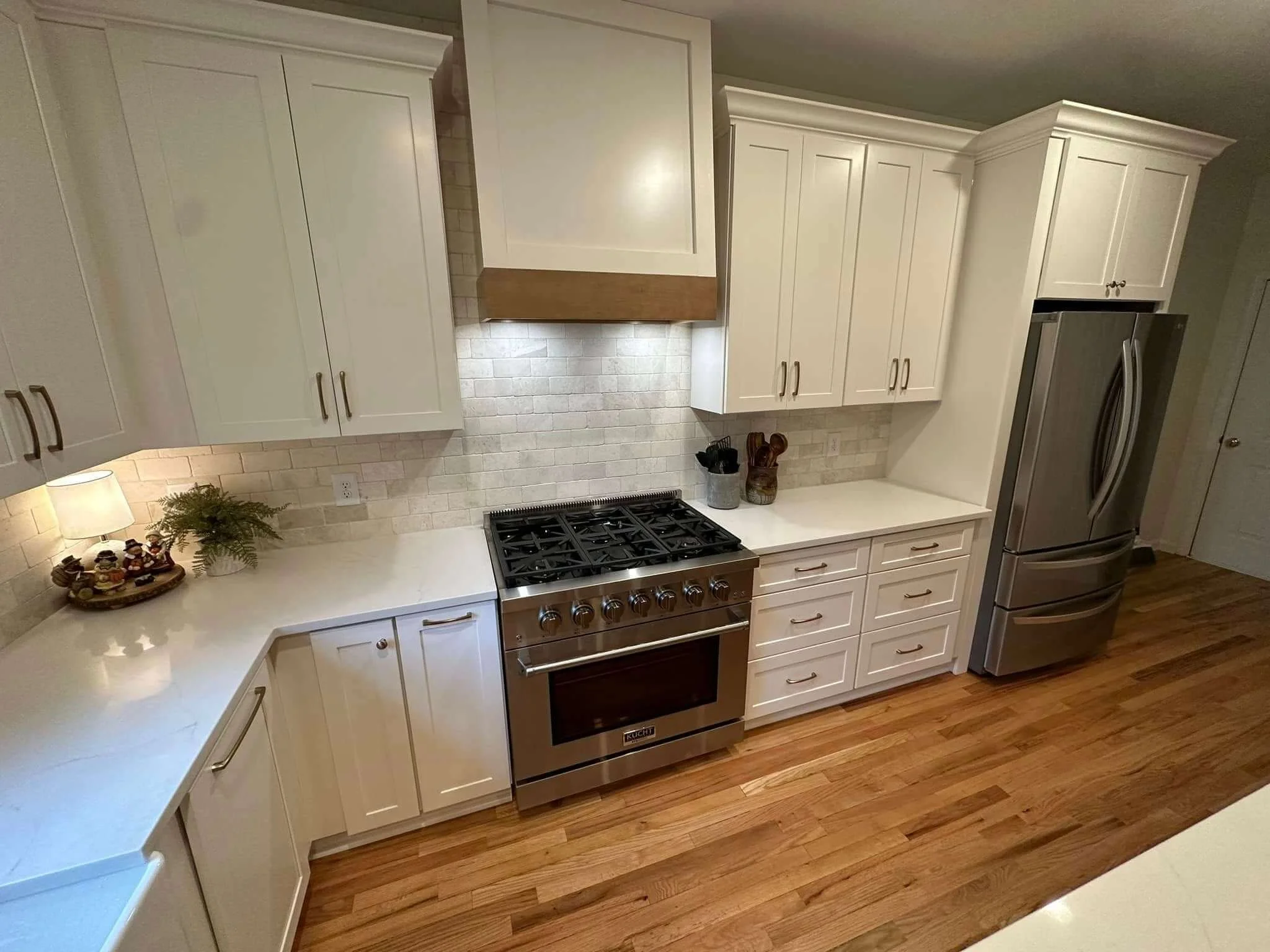 A modern kitchen with white cabinets, a stainless steel oven and refrigerator, wooden flooring, and a beige tiled backsplash.