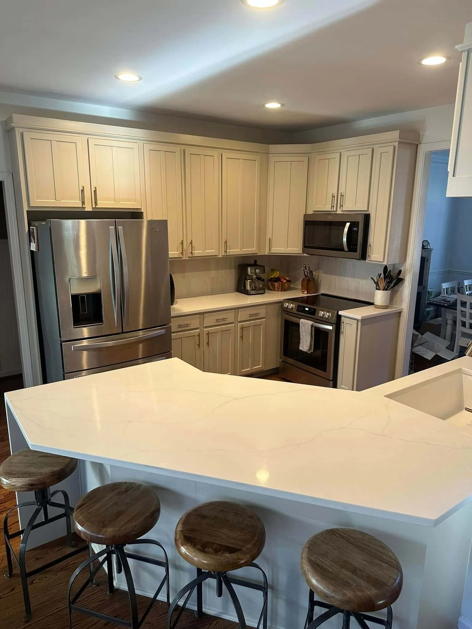 Modern kitchen with off-white cabinets, stainless steel appliances, a white island counter with four wooden stools, and cooking utensils on the counter.