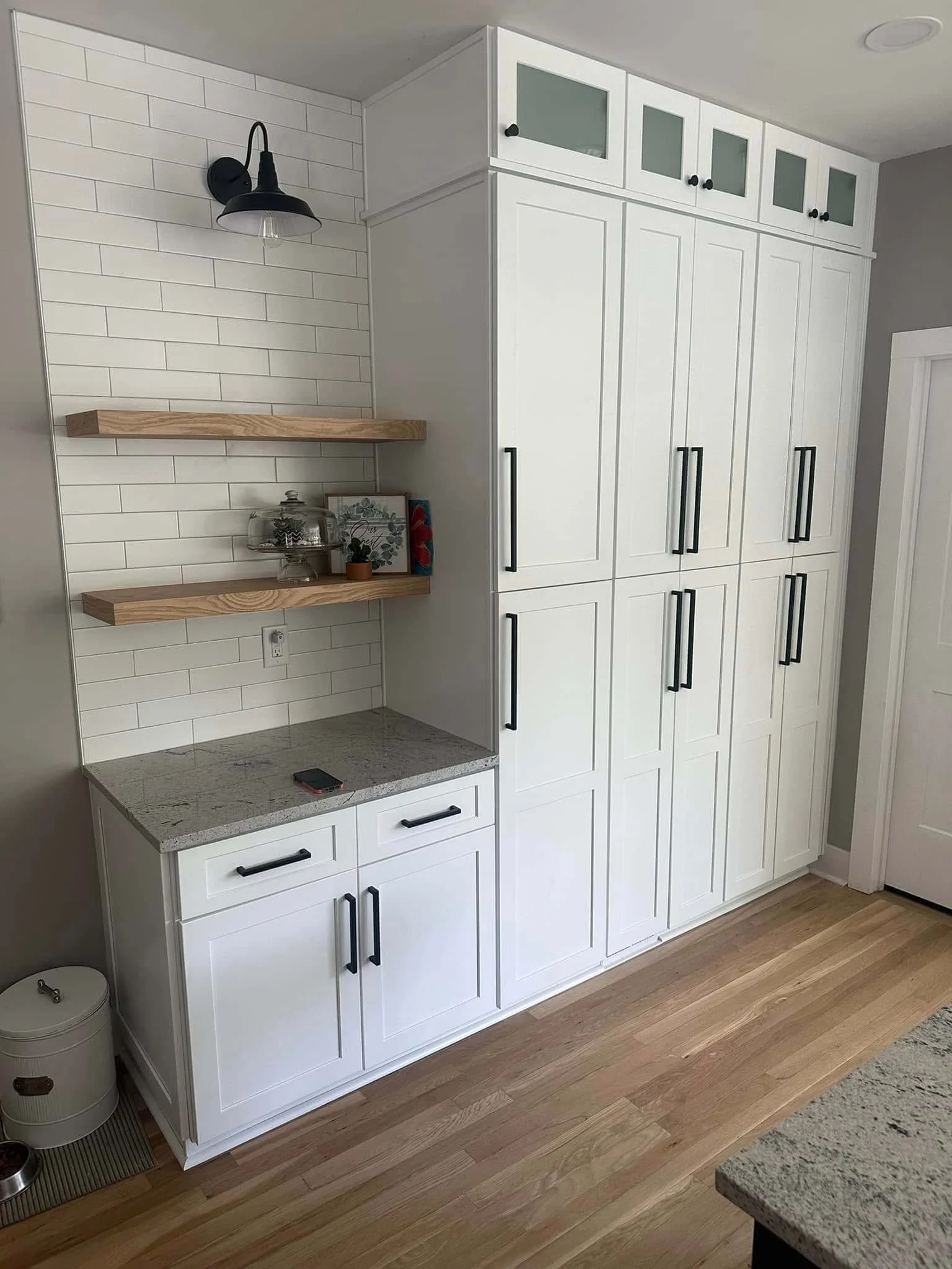 Kitchen cabinet with black handles, white tile backsplash, wooden open shelves, granite countertop, black wall-mounted lamp, and light wood flooring.