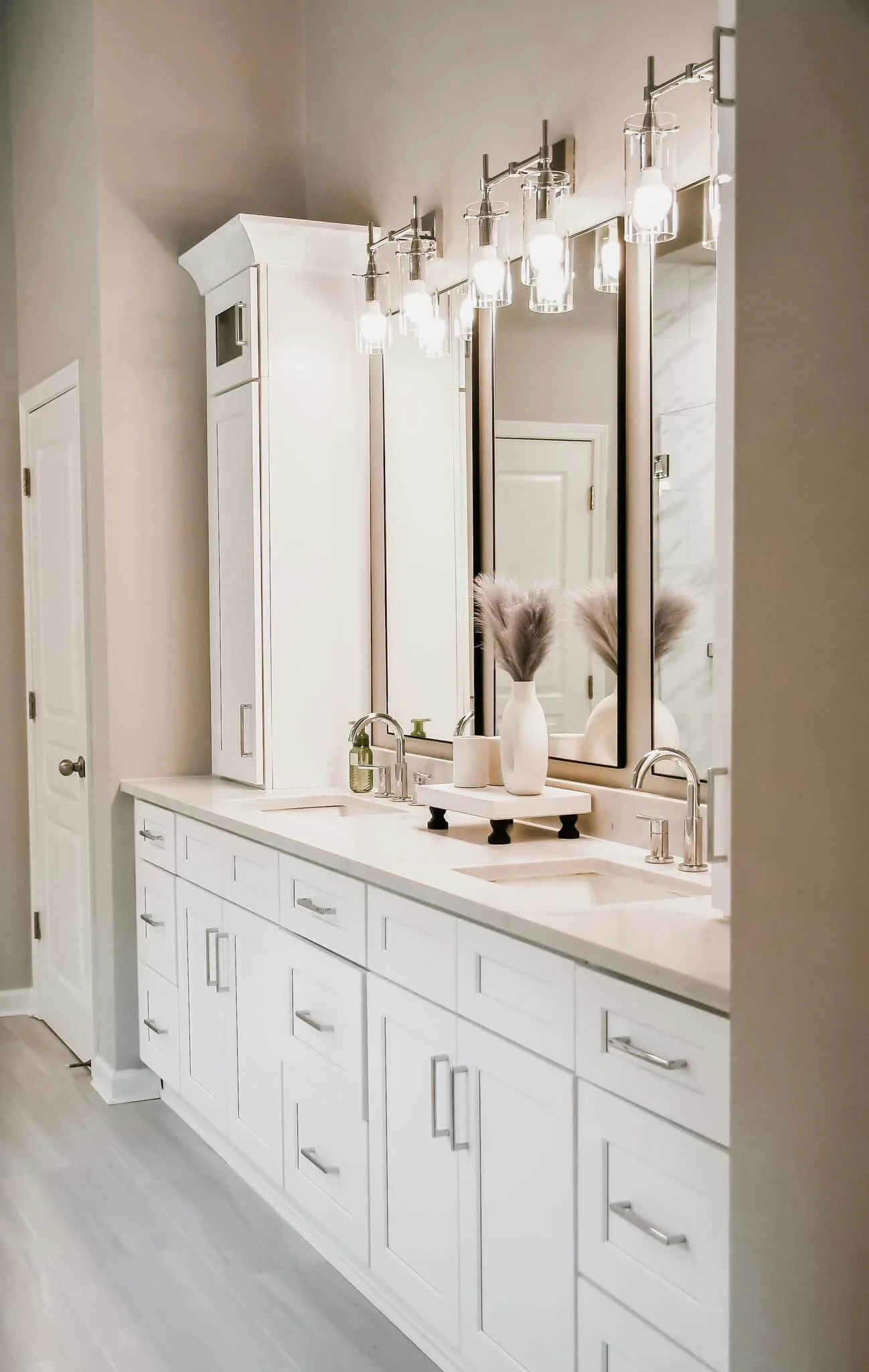 A modern bathroom vanity with dual sinks, white cabinets, large mirrors, and decorative vases with dried pampas grass, illuminated by multiple light fixtures.