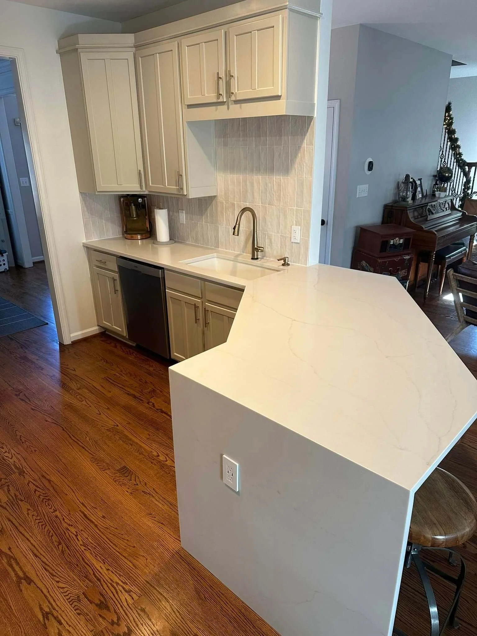 Modern kitchen with beige cabinetry, a beige countertop, a sink with a gold-colored faucet, and hardwood floors. There is a coffee maker on the counter and a decorative wall with tile backing.