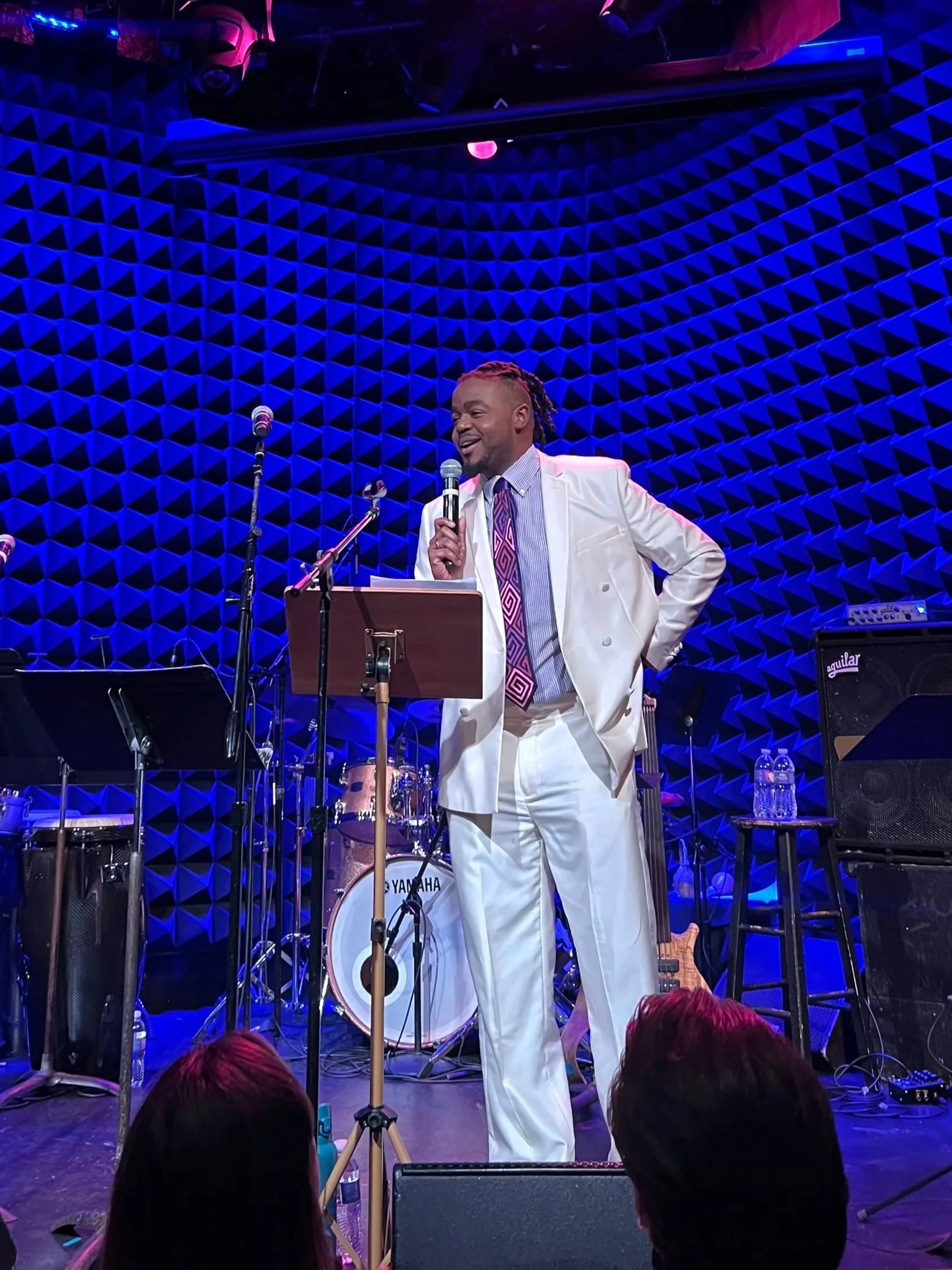 A man in a white suit holding a microphone, performing on stage with musical instruments behind him, in front of a blue geometric patterned background.
