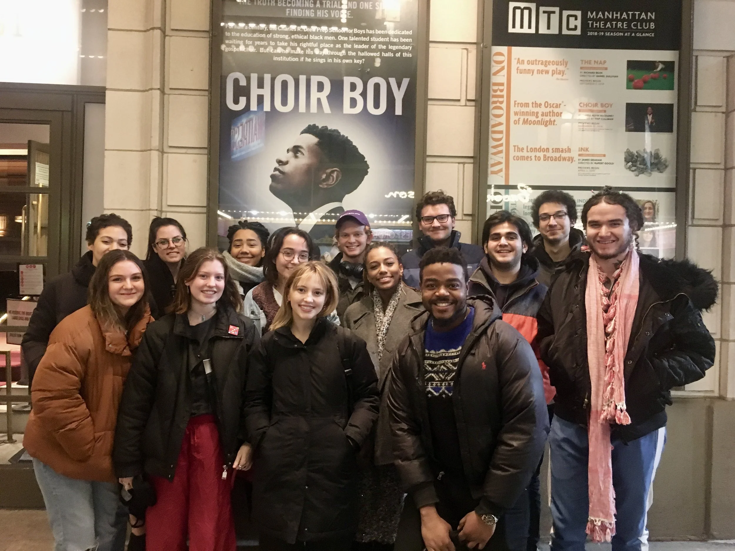 A group of fifteen young adults standing in front of a theater entrance, smiling at the camera. Behind them is a poster for the play 'Choir Boy' at the Manhattan Theatre Club, with other posters and notices on the wall.